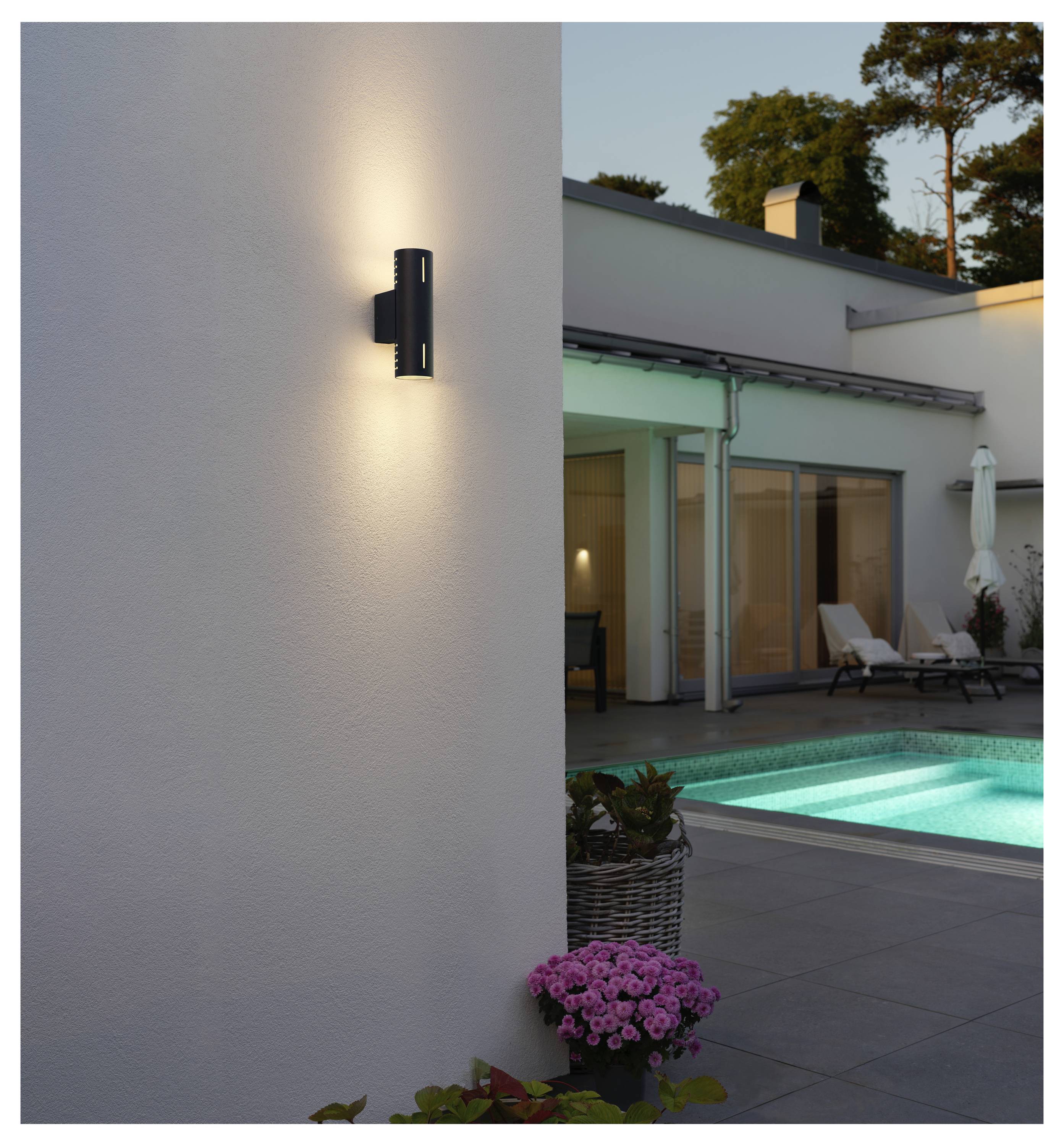 Eine moderne Außenwandleuchte beleuchtet eine Terrassenanlage neben einem Pool, wobei im Hintergrund bei Einbruch der Dämmerung ein zeitgenössisches Haus zu sehen ist.