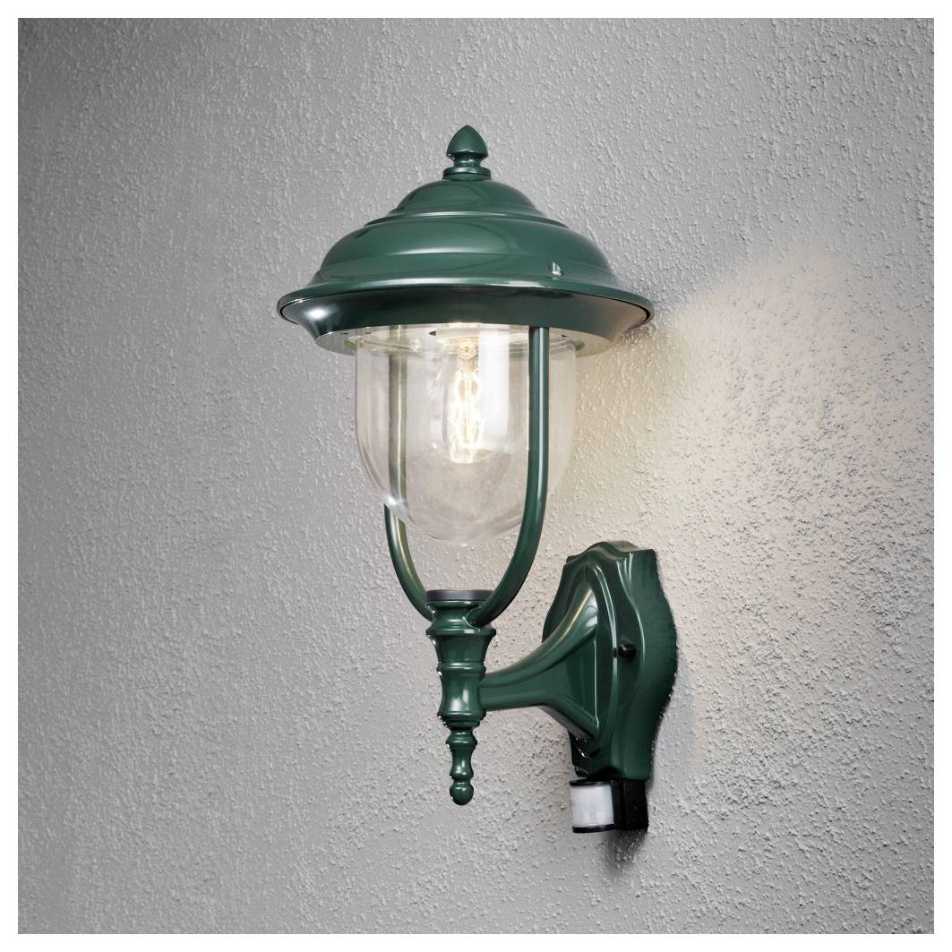 Eine Wandlaterne im Vintage-Stil in Grün mit einer klaren Glühbirne, die warmes Licht vor einer strukturierten grauen Wand ausstrahlt.