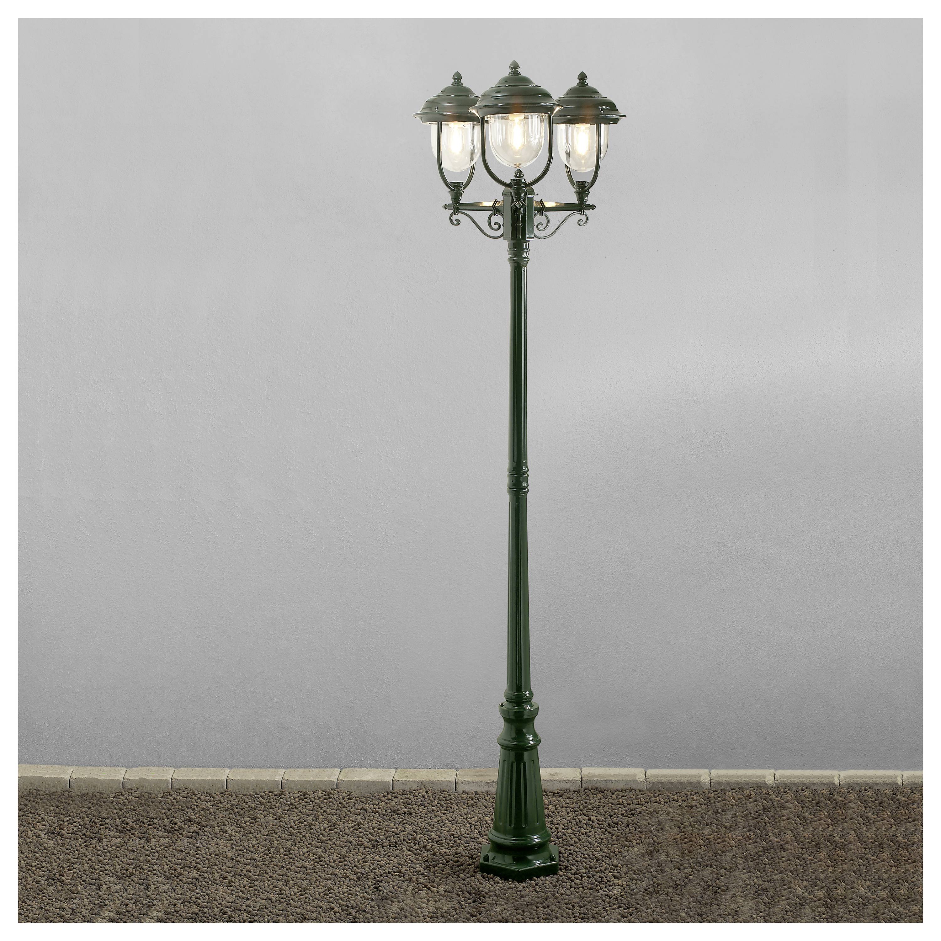 Eine im Vintage-Stil gehaltene Straßenlaterne mit drei Laternen, deren jede kunstvoll gearbeitetes Metallwerk aufweist, steht auf einem Kiesweg vor einem schlichten, nebligen Hintergrund.