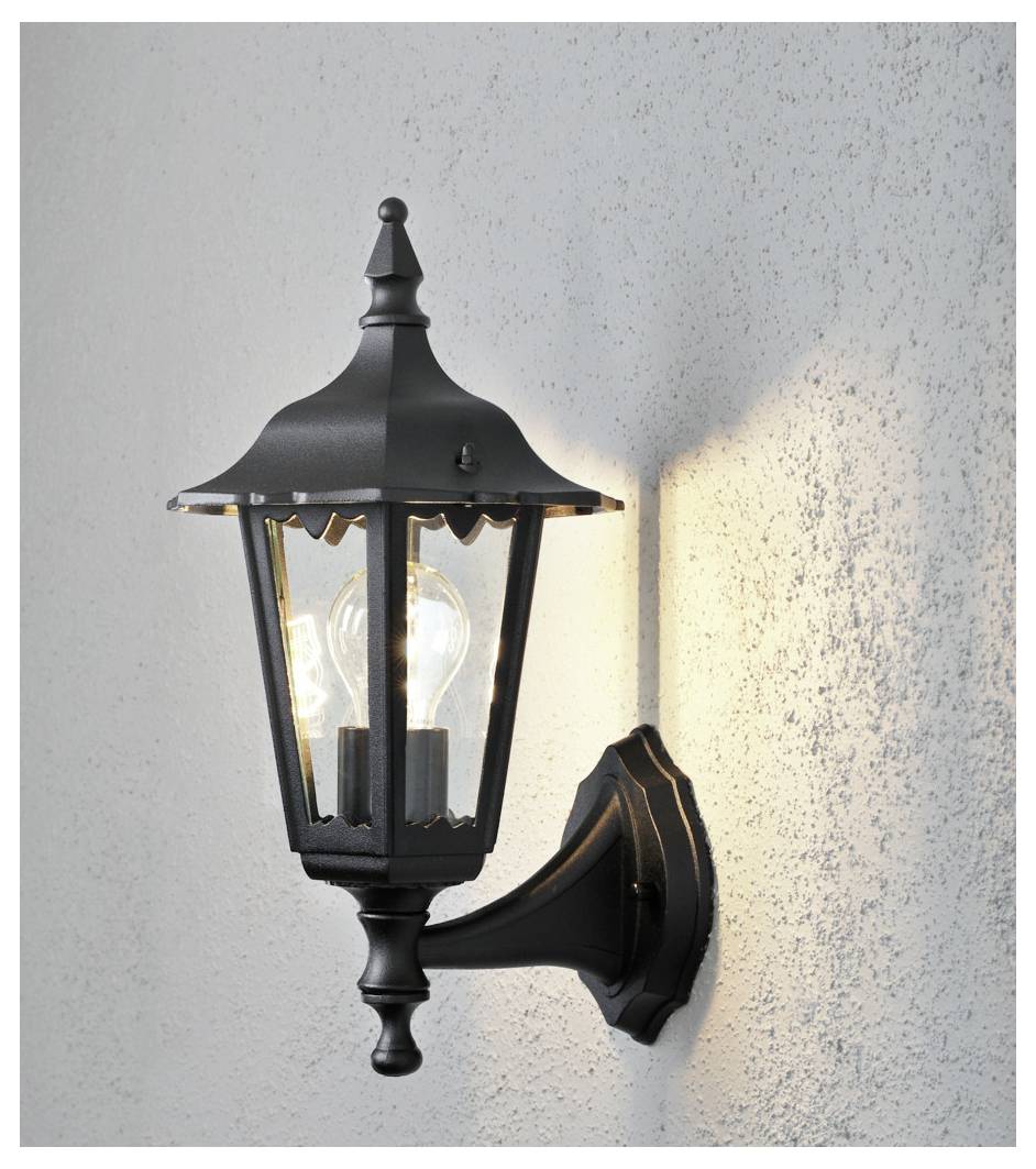 Eine schwarze, verzierte Wandlaterne im Außenbereich mit einer leuchtenden Glühbirne, die Licht auf eine strukturierte graue Wand wirft.