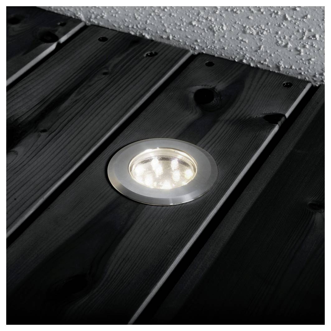 Ein rundes, eingelassenes Bodenleuchte ist in ein dunkles Holzdeck eingebettet und wirft Licht auf die Dielen, die parallel zu einer hellgefärbten Wand ausgerichtet sind.