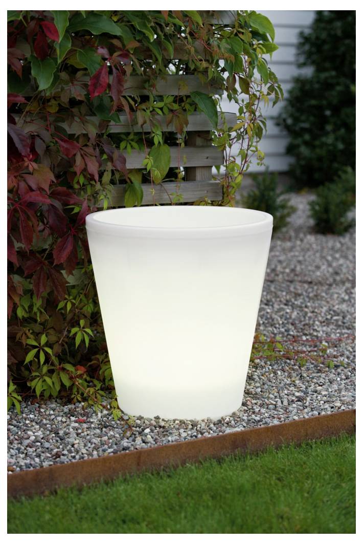 Ein leuchtend weißer Pflanzenkübel auf einer Kiesoberfläche neben grüner und roter Vegetation, mit Gras im Vordergrund und einem Haushindergrund.