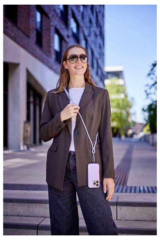 Eine Frau steht im Freien und lächelt, trägt eine Sonnenbrille und eine Jacke, hält ein Telefon mit einem Riemen um den Hals.