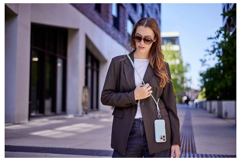 Eine Person geht auf einer Stadtstraße im Freien, trägt eine Sonnenbrille und einen Blazer und hält ein Smartphone an einem Gurt um den Hals.