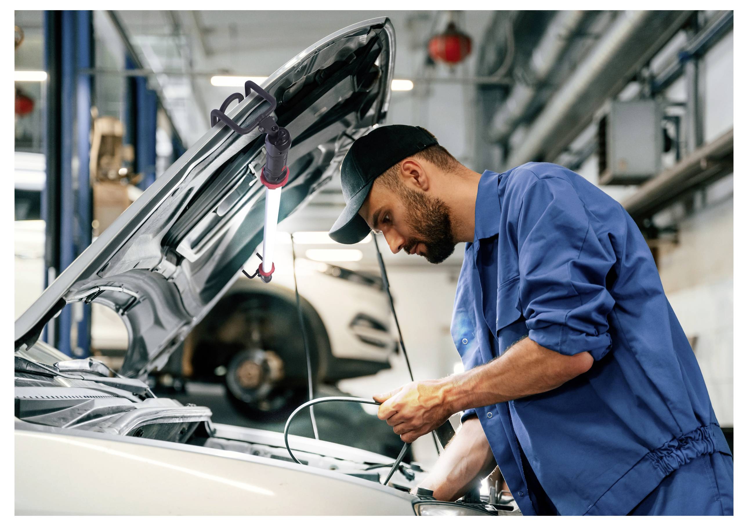 Ein Mechaniker in blauer Uniform arbeitet an einem Automotor in einer Werkstatt, mit offener Motorhaube und Werkzeugen in der Hand, konzentriert auf die Reparaturen fokussierend.