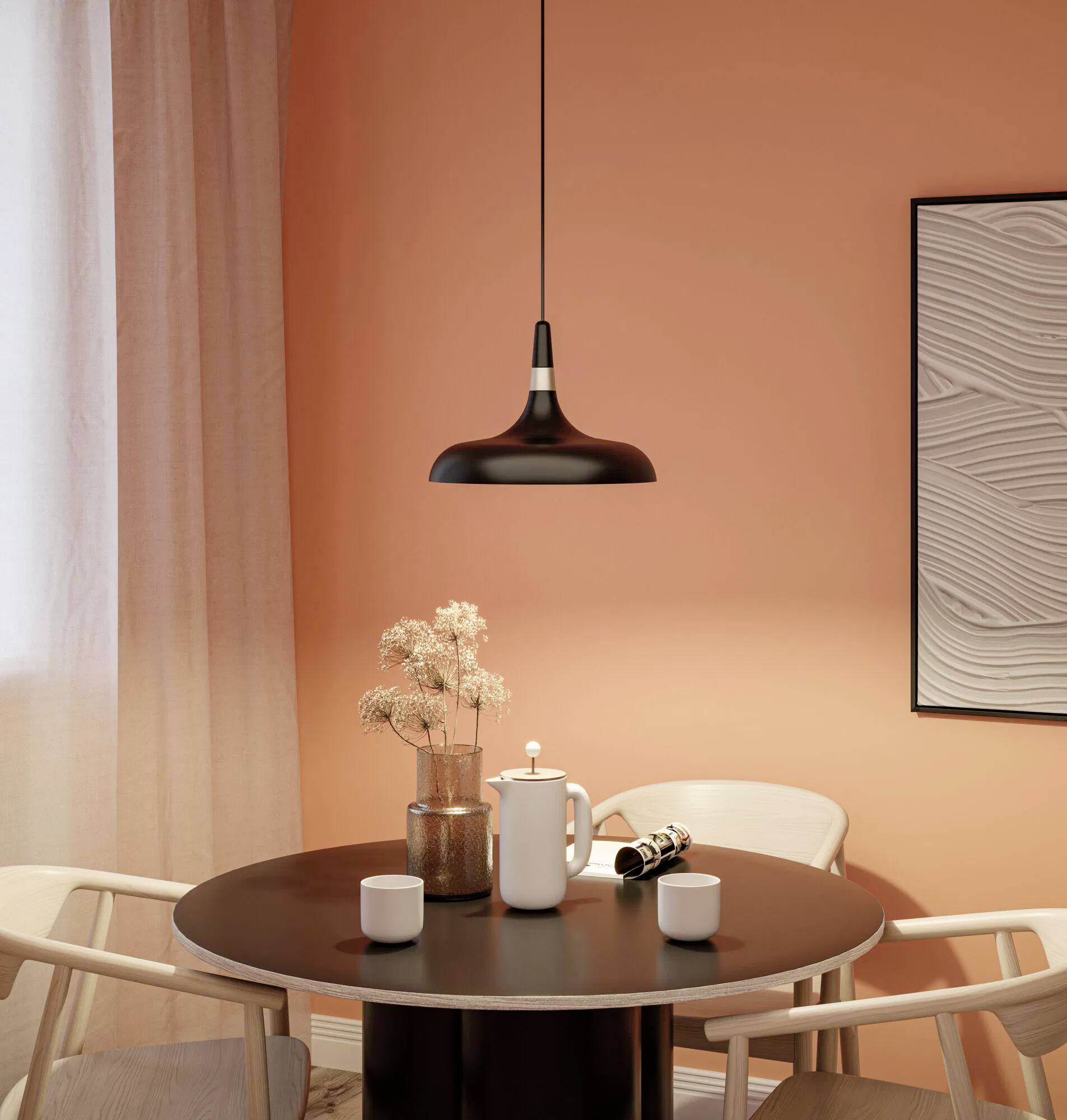 Ein gemütlicher Essbereich mit einer orangefarbenen Wand zeigt einen runden Tisch, zwei Tassen, eine Teekanne und getrocknete Blumen unter einer Pendelleuchte.