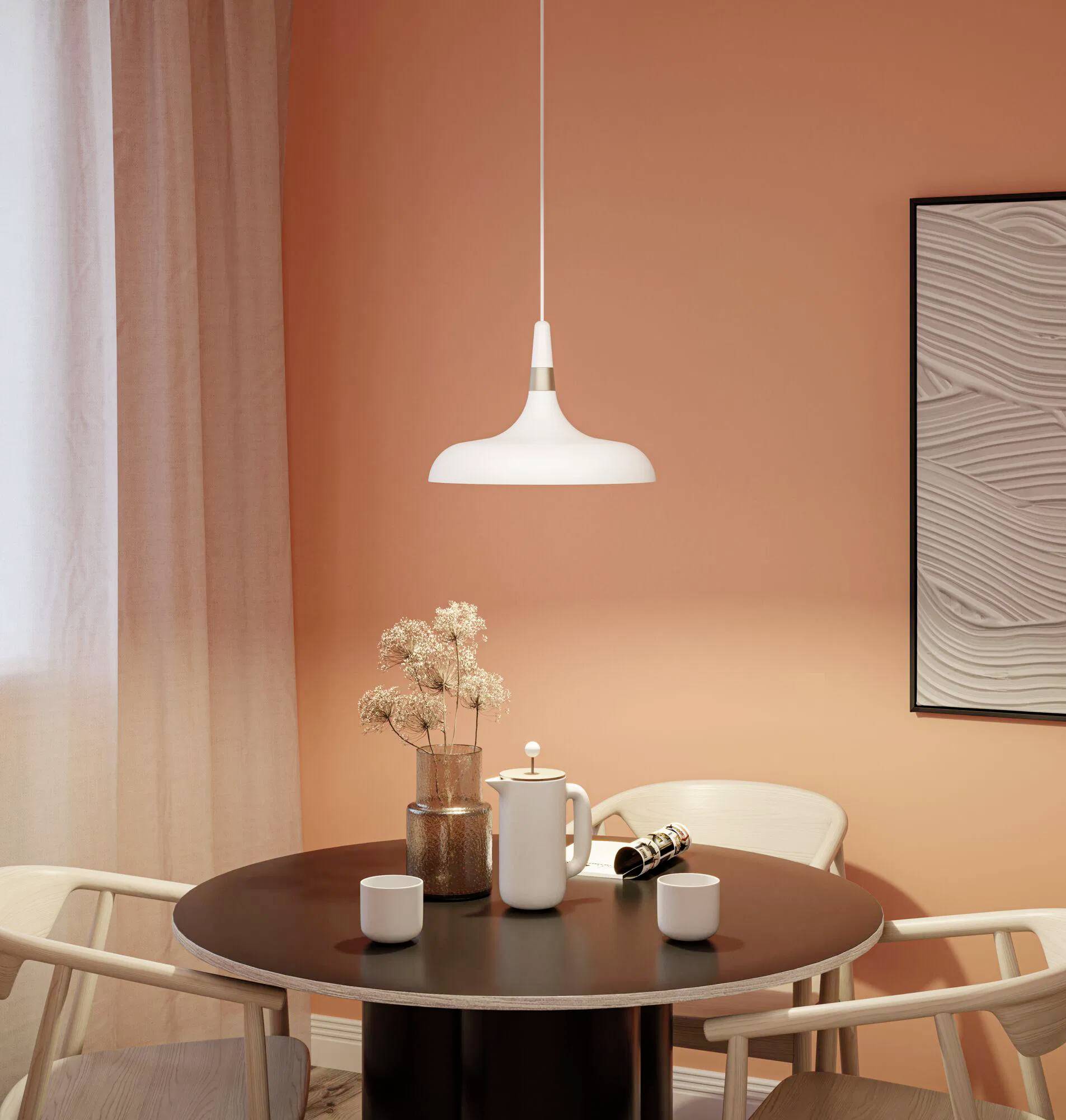 Ein kleiner runder Esstisch mit zwei Stühlen, einer Kaffeekanne, Tassen und getrockneten Blumen unter einer weißen Hängelampe vor einer orangefarbenen Wand.