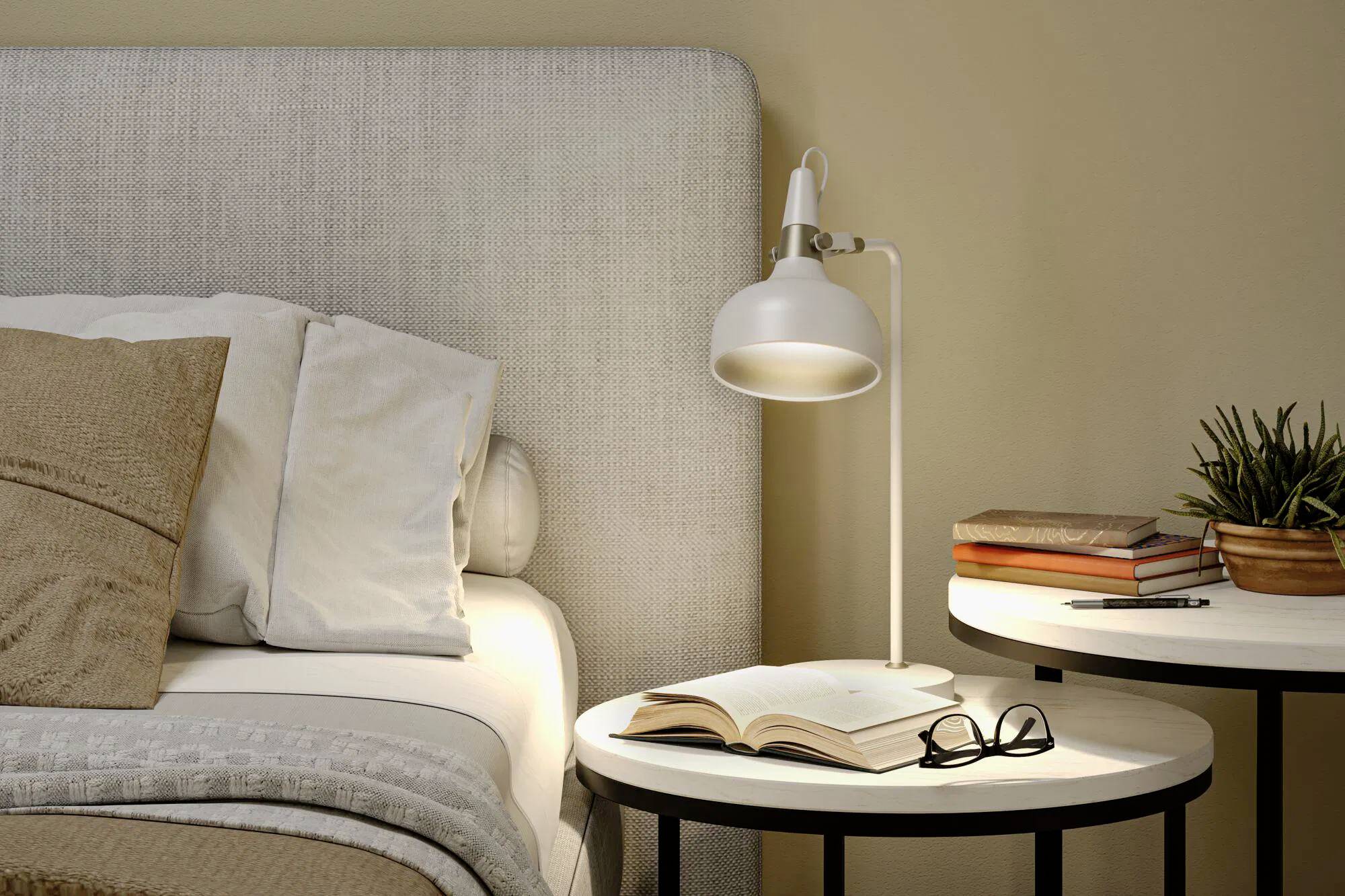 Eine gemütliche Schlafzimmerszene mit einem Bett, einer beleuchteten Lampe auf einem Nachttisch, einem aufgeschlagenen Buch und einer Brille. Auf einem weiteren Tisch stehen Bücher und eine Topfpflanze.