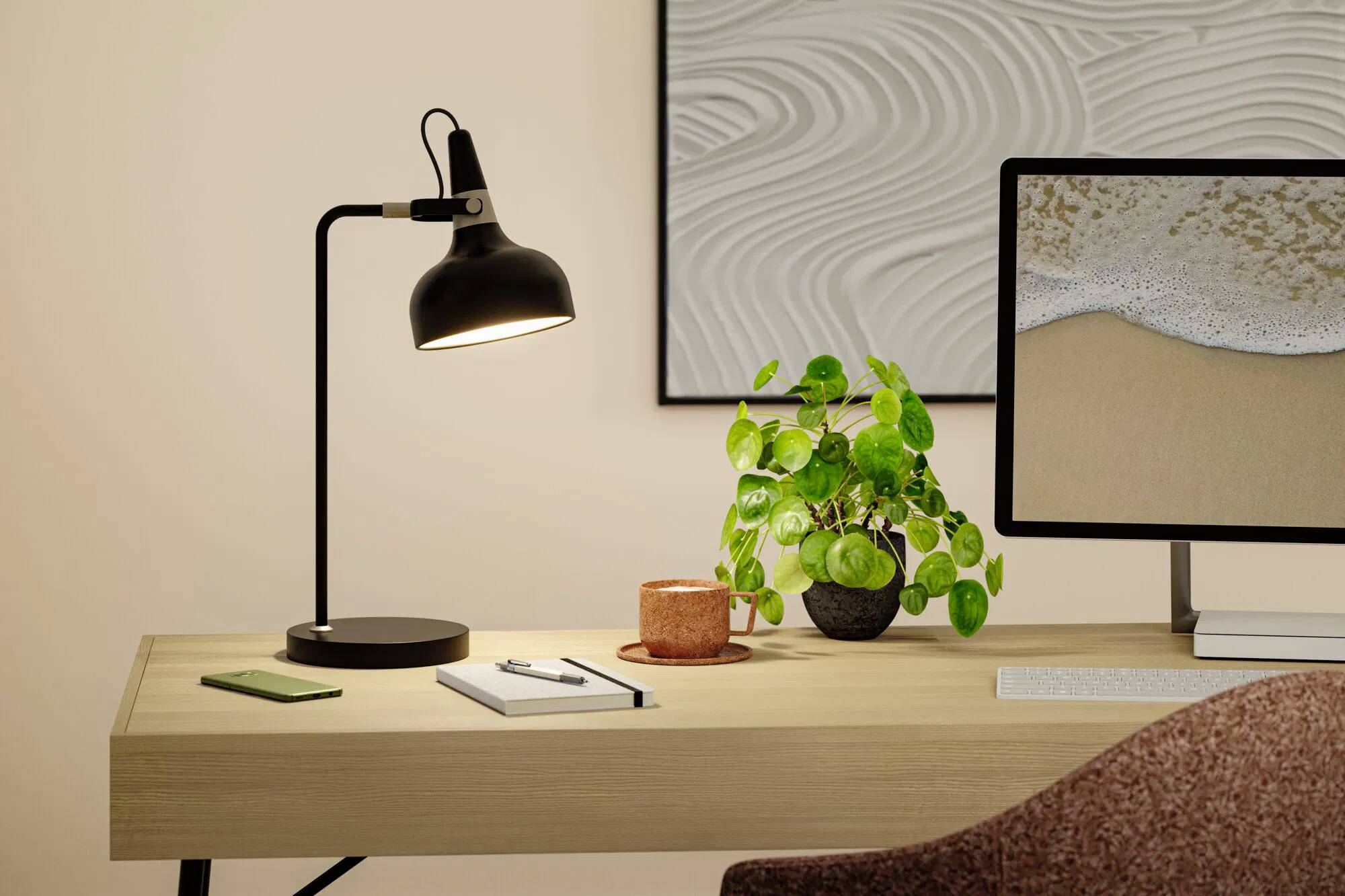 Ein modernes Schreibtisch-Setup mit einer schwarzen Lampe, einer Topfpflanze, einem Notizbuch und einem Smartphone. Ein Computer zeigt ein Strandbild an. Kunst schmückt die Wand.