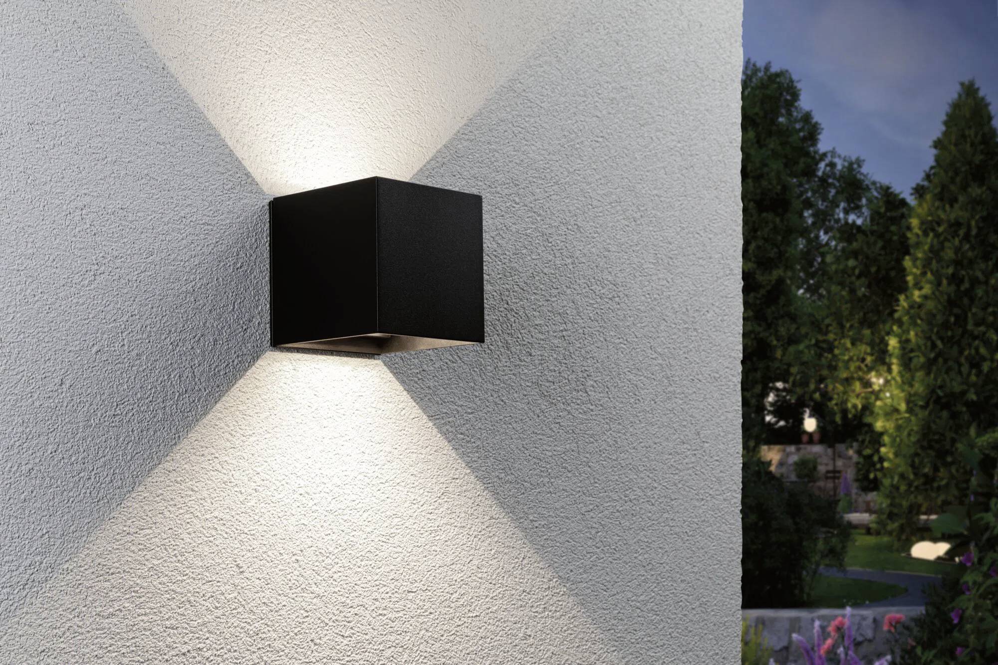 Eine moderne schwarze Wandleuchte an einer strukturierten weißen Außenwand, die symmetrische Lichtmuster wirft. Garten und Bäume sind im Hintergrund sichtbar.