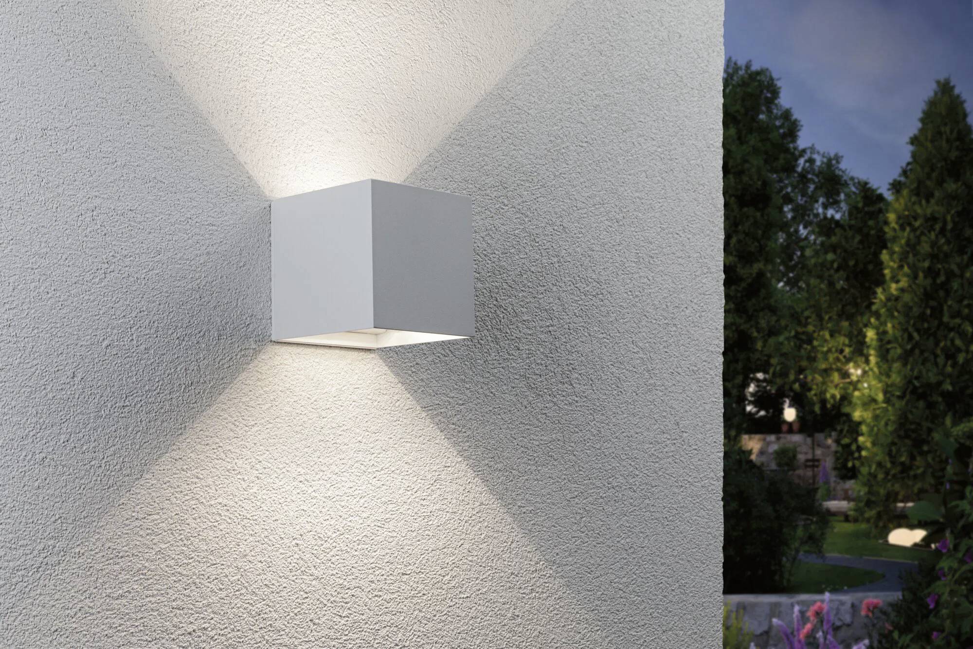 Wandmontierte Würfelleuchte an einer strukturierten Außenwand, die symmetrische Schatten in V-Form wirft. Bäume im Hintergrund.