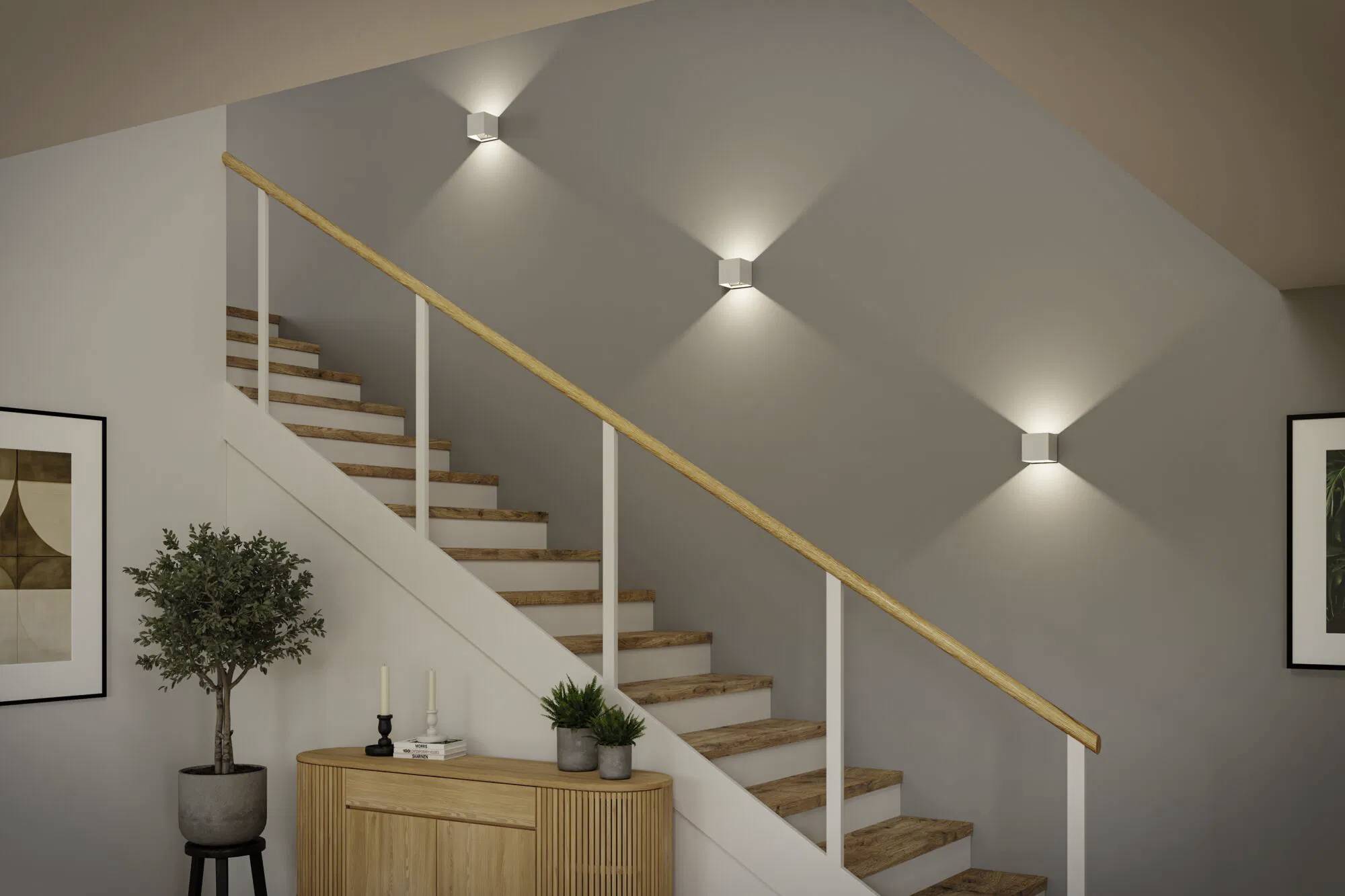Eine Holztreppe mit einem Handlauf wird von drei wandmontierte Leuchten beleuchtet. An der Basis steht ein kleiner Schrank mit Pflanzen und Kerzen.