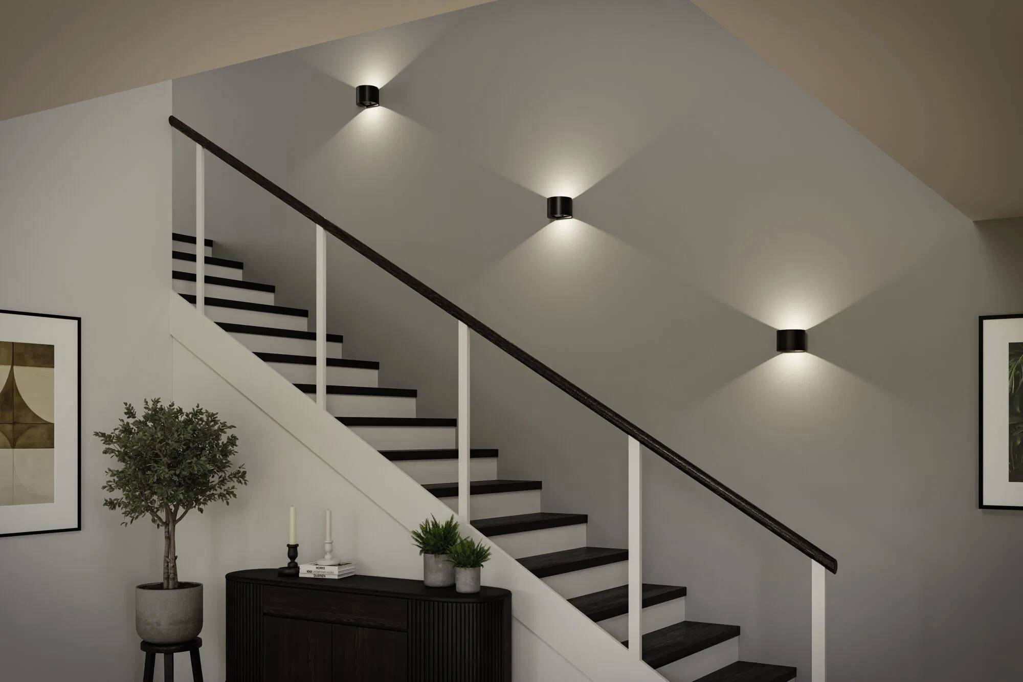 Eine moderne, gut beleuchtete Treppe mit schwarzen Stufen, weißen Geländern und wandmontierten Lichtarmaturen. Ein kleiner Tisch mit Pflanzen und gerahmter Kunst in der Nähe.