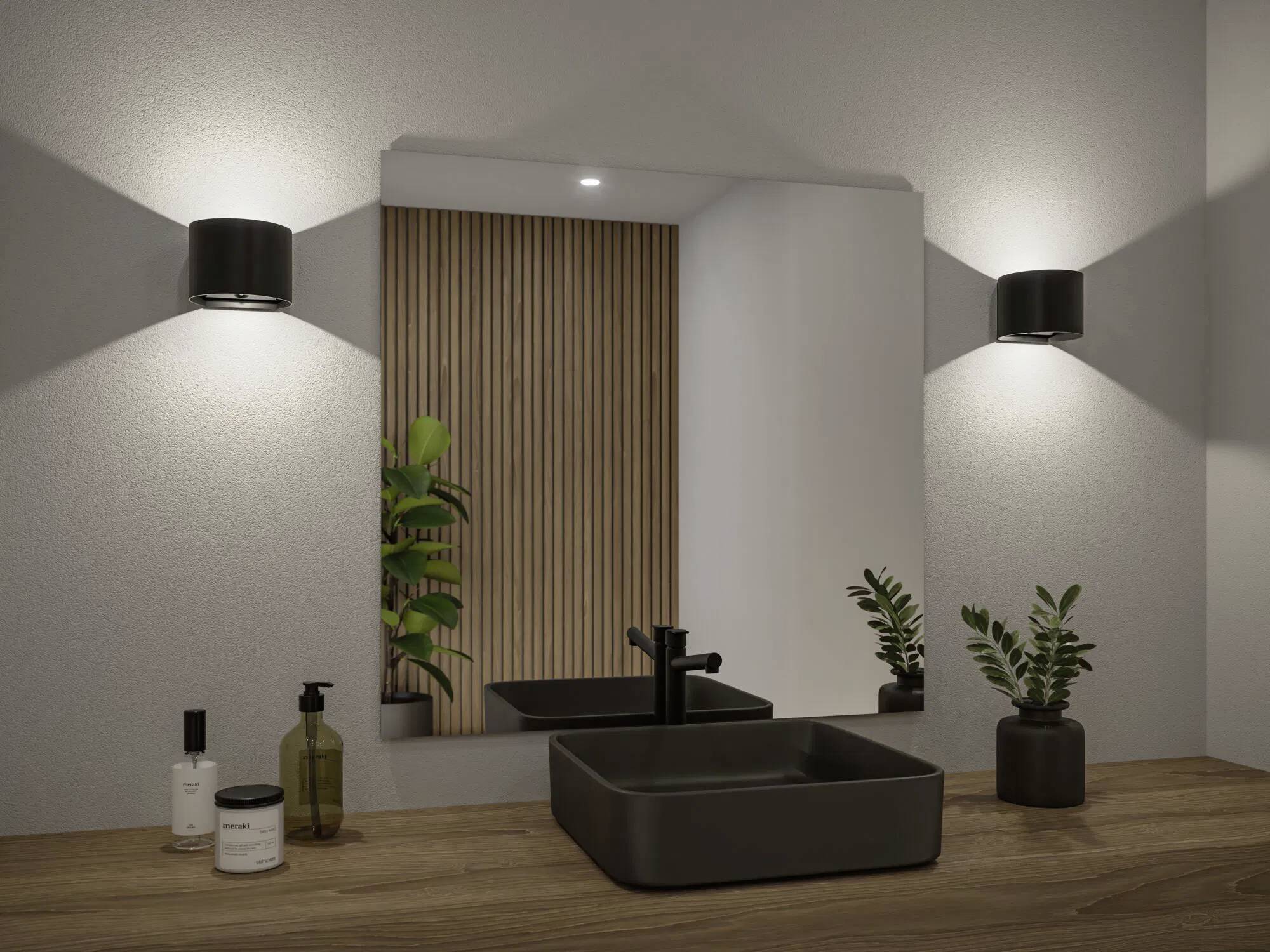 Modernes Badezimmer-Waschtisch mit rechteckigem Spiegel, Holzarbeitsplatte, schwarzem Waschbecken, Wasserhahn und Topfpflanzen. Wandleuchten werfen ein weiches Licht.