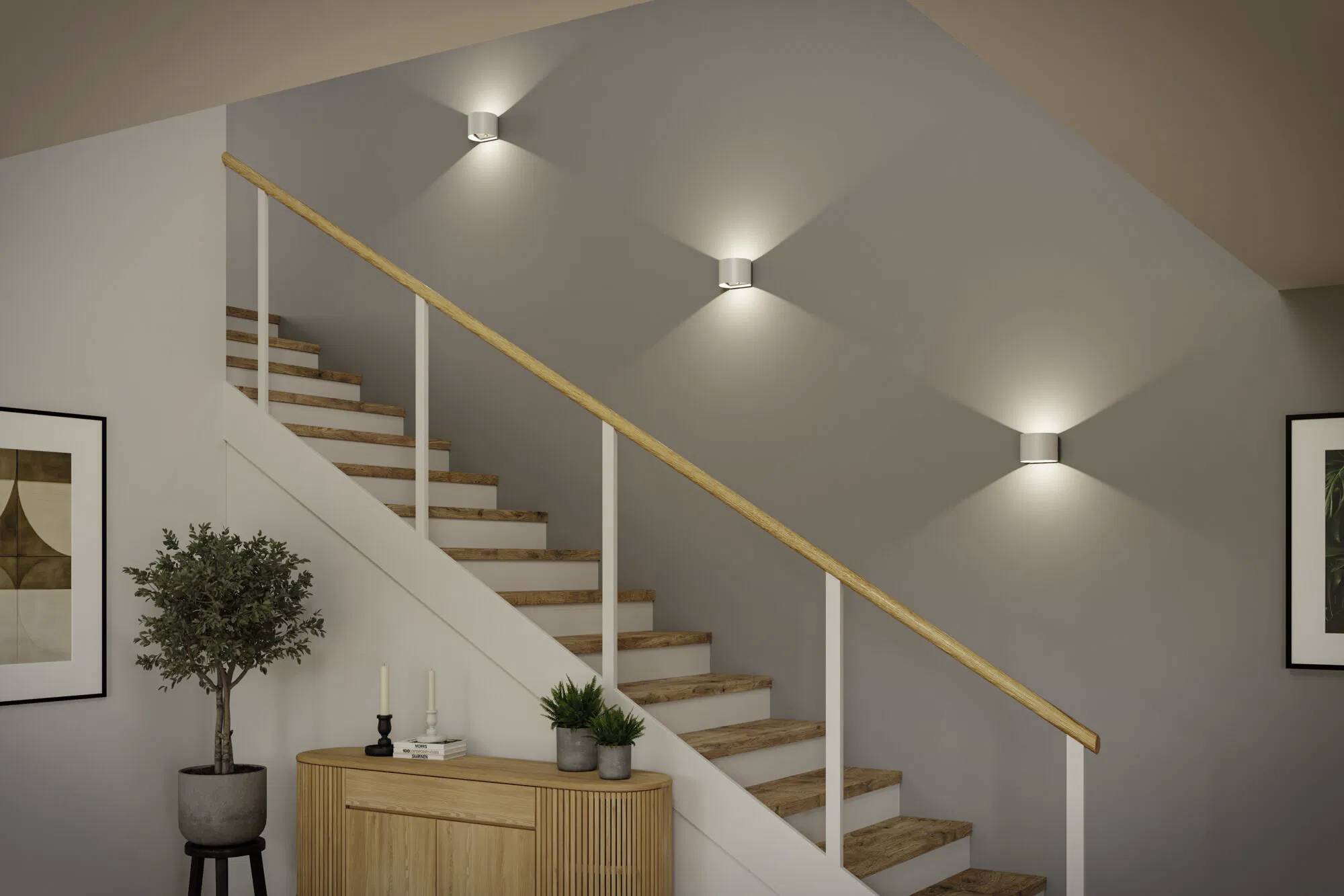 Eine moderne Holztreppe mit einem hellbraunen Handlauf und wandmontierter Beleuchtung. Ein kleiner Tisch mit Pflanzen und Dekorationen steht darunter.