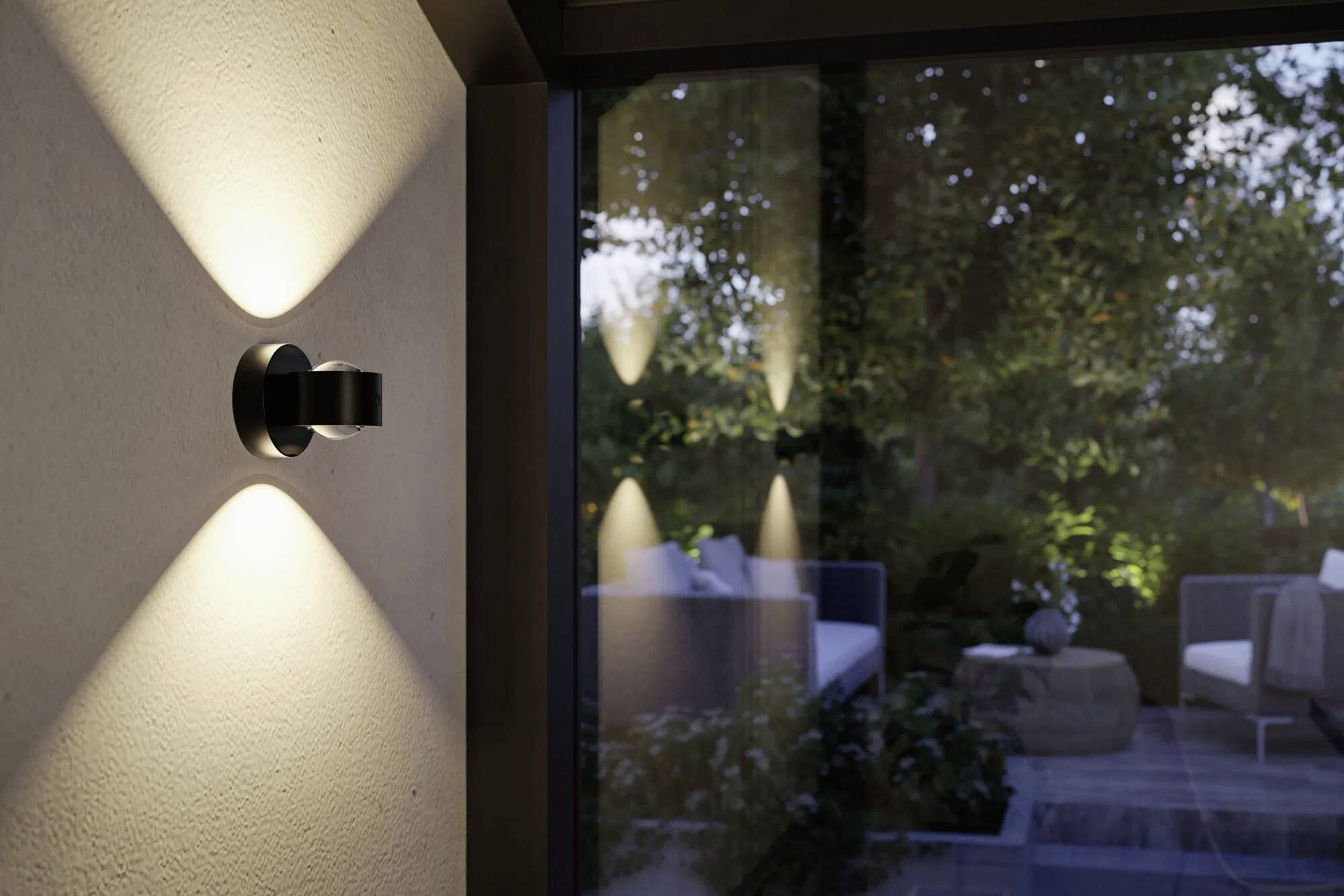 Wandlampe, die eine Terrasse bei Einbruch der Dämmerung beleuchtet, mit Außensitzgelegenheiten, die durch eine Glastür sichtbar sind, umgeben von Grünpflanzen und sanfter Beleuchtung.