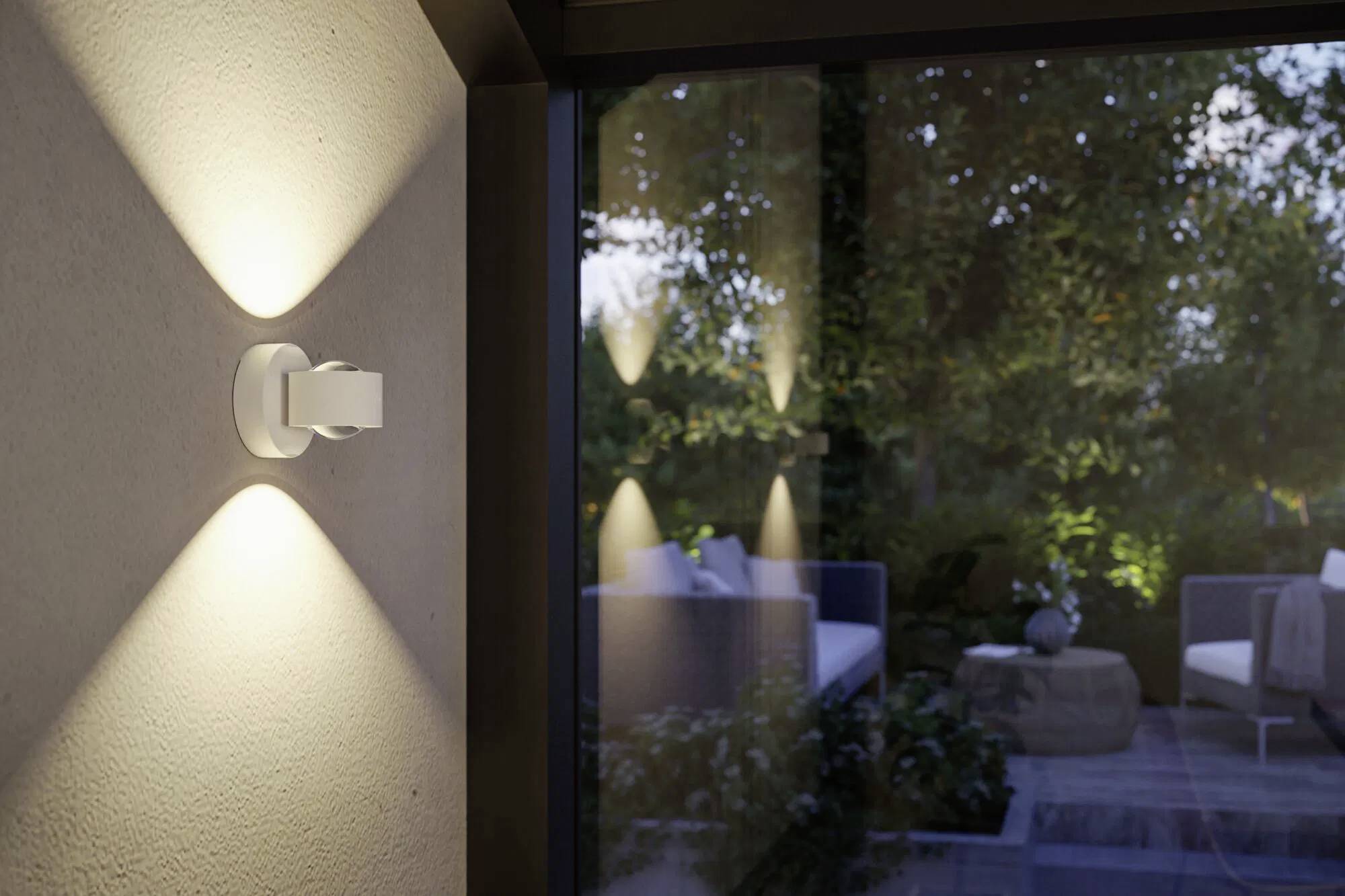 Eine moderne wandmontierte Leuchte strahlt auf einer Außenterrasse und beleuchtet Sitzmöbel und Grünpflanzen.