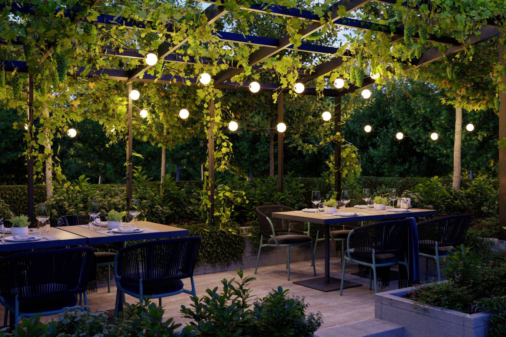 Outdoor-Restaurantbereich mit Tischen und Stühlen unter einer Pergola, die mit Lichterketten und üppiger Grünpflanzung geschmückt ist und eine einladende Atmosphäre schafft.