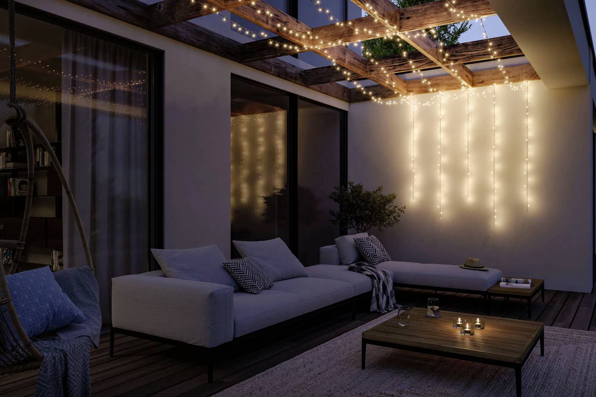 Überdachte Terrasse, beleuchtet durch hängende Lichterketten und ein warmes, deckenartiges Leuchten. Der Sitzbereich umfasst Kissen und einen Couchtisch.