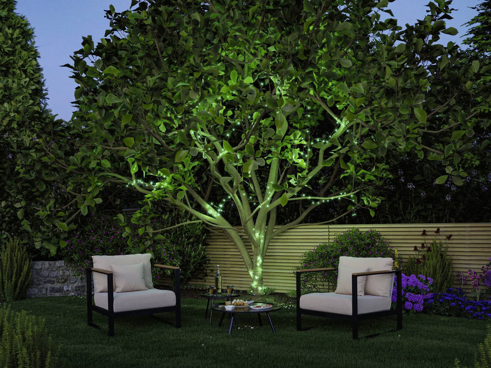 Gartenszene bei Einbruch der Dämmerung mit einem Baum, der von grünem Licht beleuchtet wird. Zwei beige Stühle stehen sich gegenüber an einem kleinen Tisch, auf dem Snacks und eine Kerze stehen.