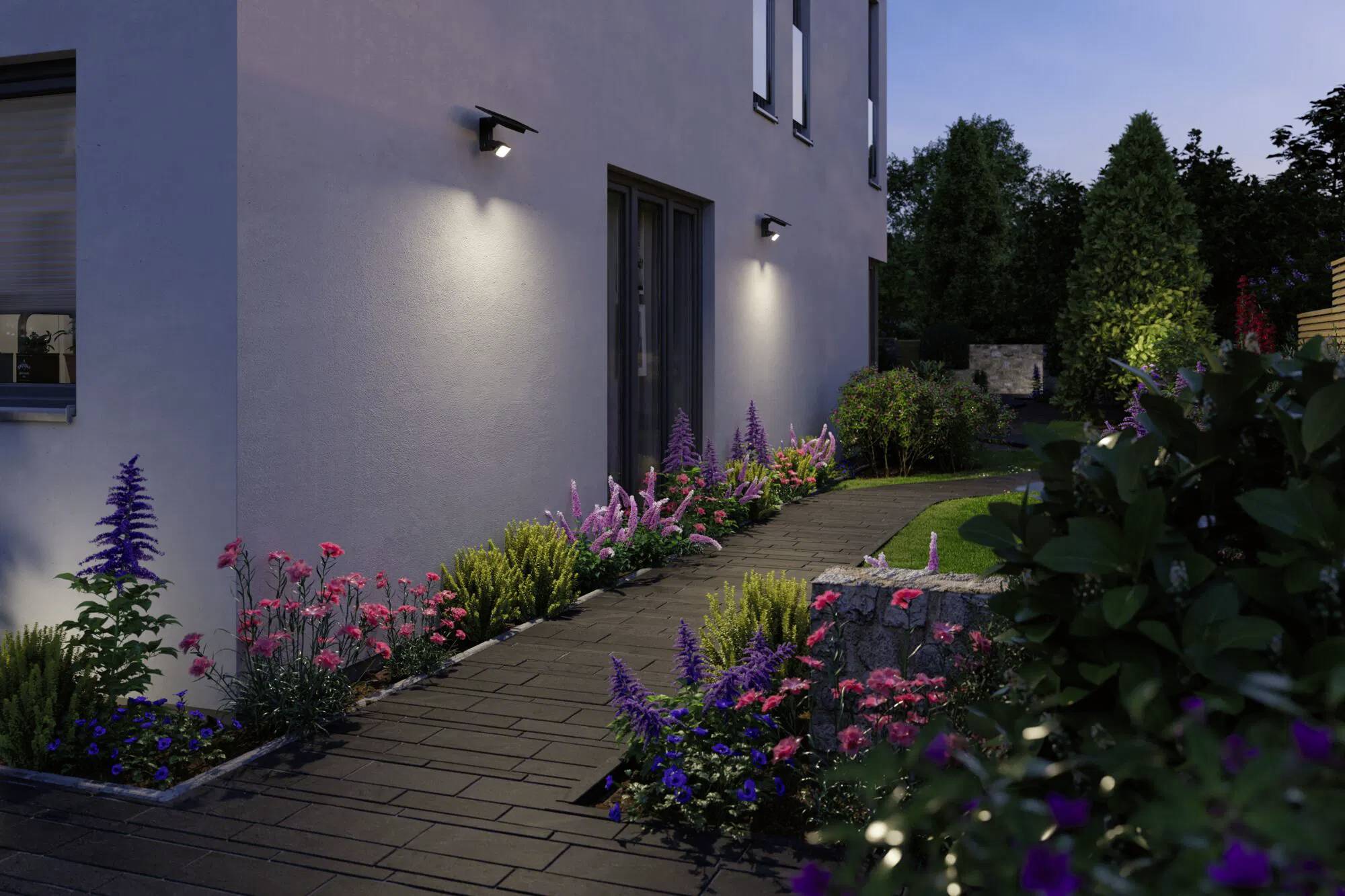 Weg mit bunten Blumen und Sträuchern, der zum Hauseingang führt und am Abend durch Außenwandleuchten beleuchtet wird.