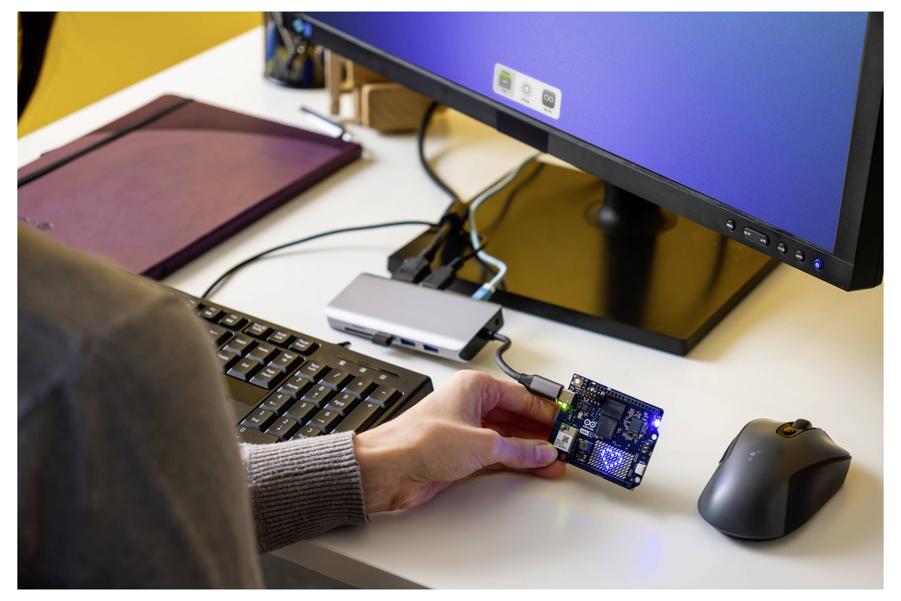 Eine Person hält eine kleine Leiterplatte, die mit einem Computer verbunden ist und wahrscheinlich für Programmierung oder Hardware-Tests verwendet wird, auf einem Schreibtisch mit Tastatur und Maus.