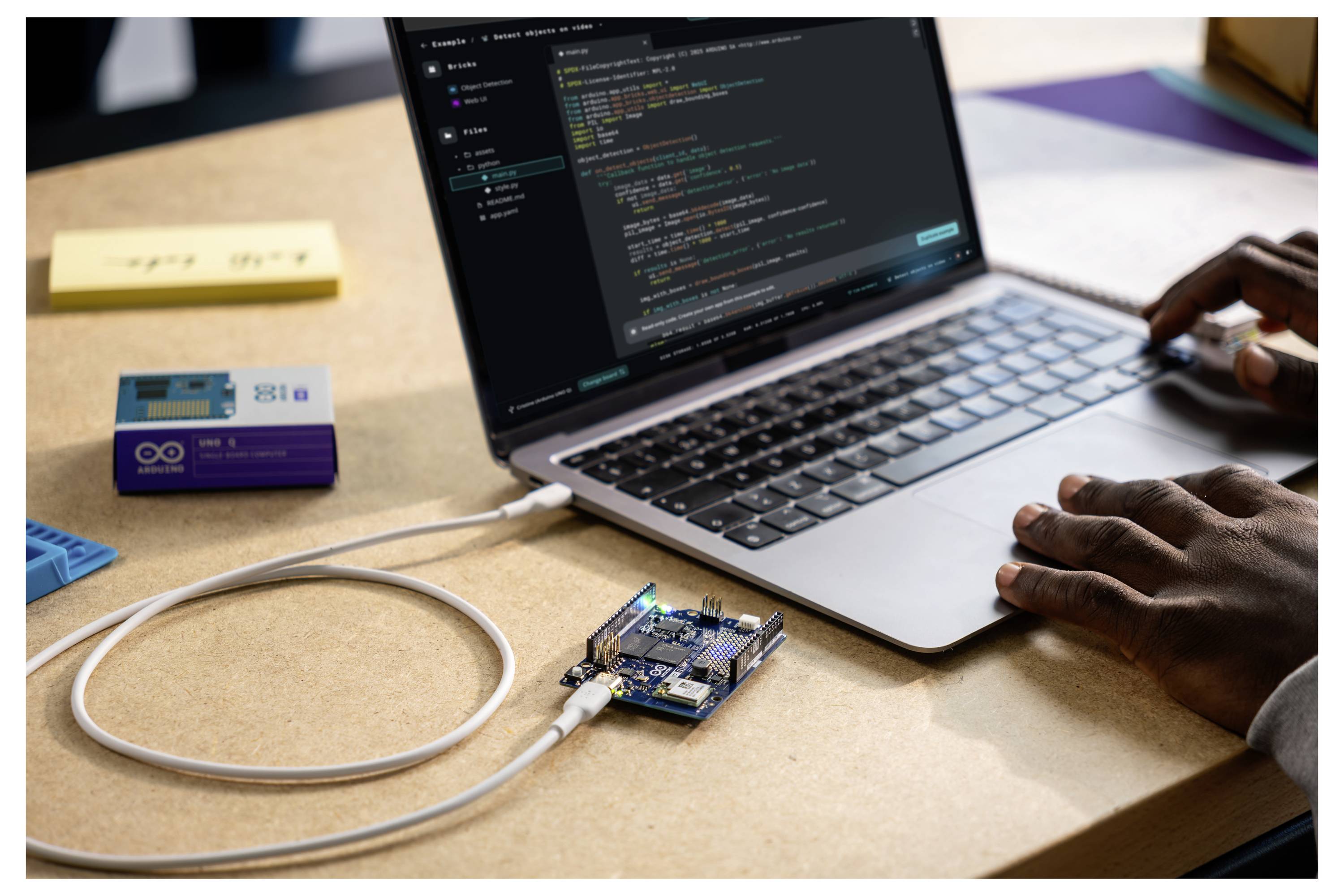 Eine Person tippt auf einem Laptop, der über ein USB-Kabel mit einem Mikrocontroller verbunden ist. Die Coding-Software ist geöffnet, und daneben liegt eine kleine Box.