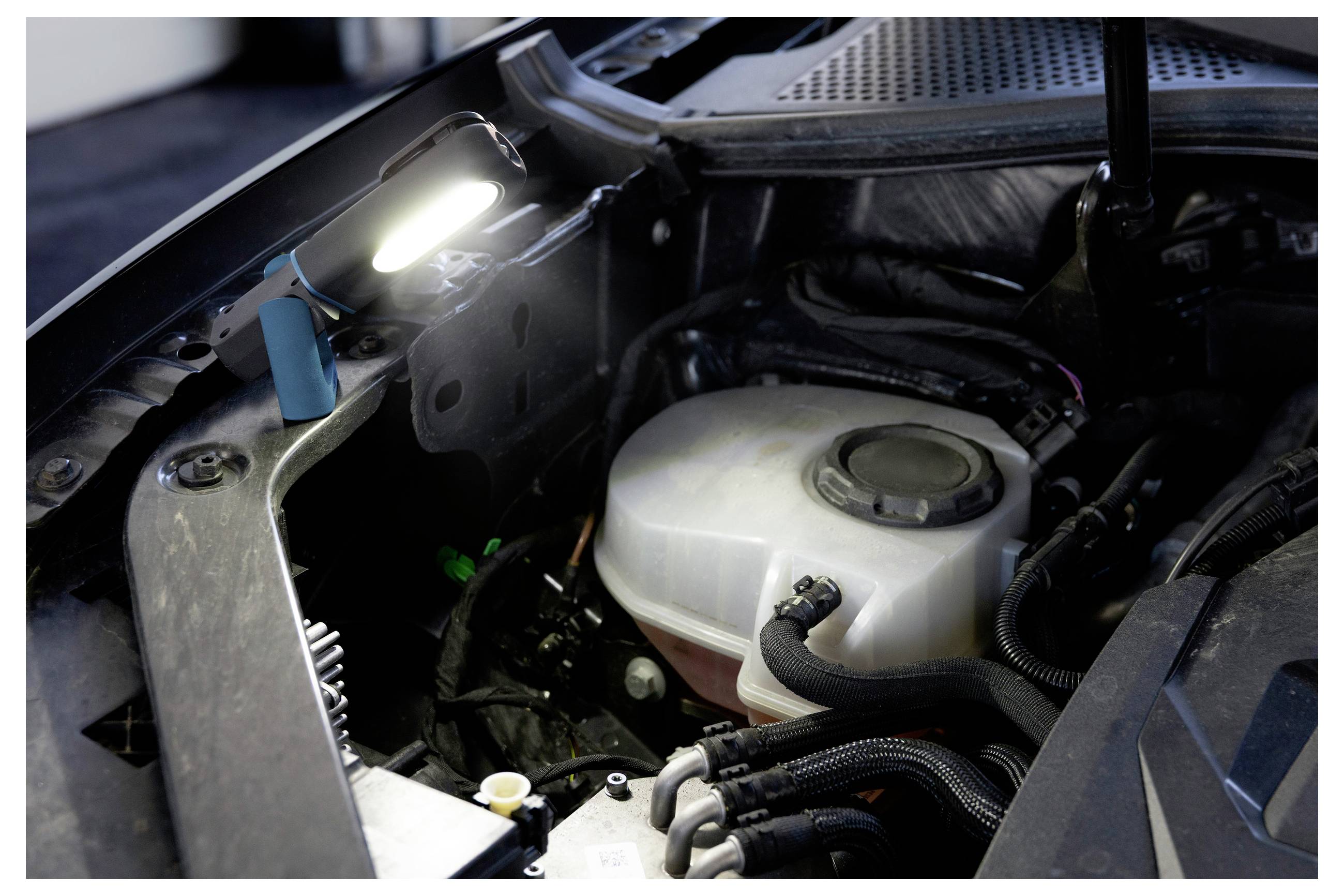 Ein beleuchteter Motorraum zeigt einen Kühlmittelbehälter und verschiedene Schläuche, wobei eine Lampe den Bereich für Wartungsarbeiten beleuchtet.