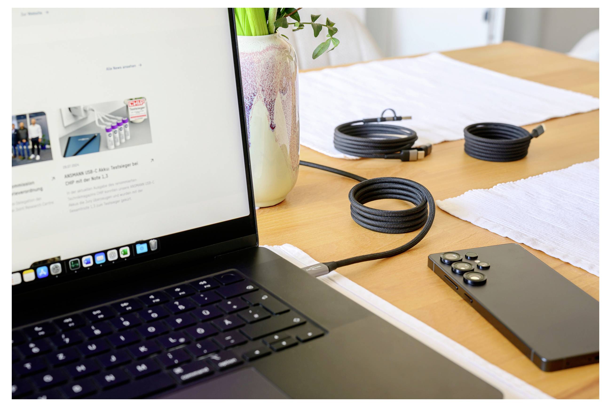 Ein Laptop auf einem Holztisch neben einem Smartphone und schwarzen Kabeln, in der Nähe einer Vase mit grünen Pflanzen, in einem hellen Raum.