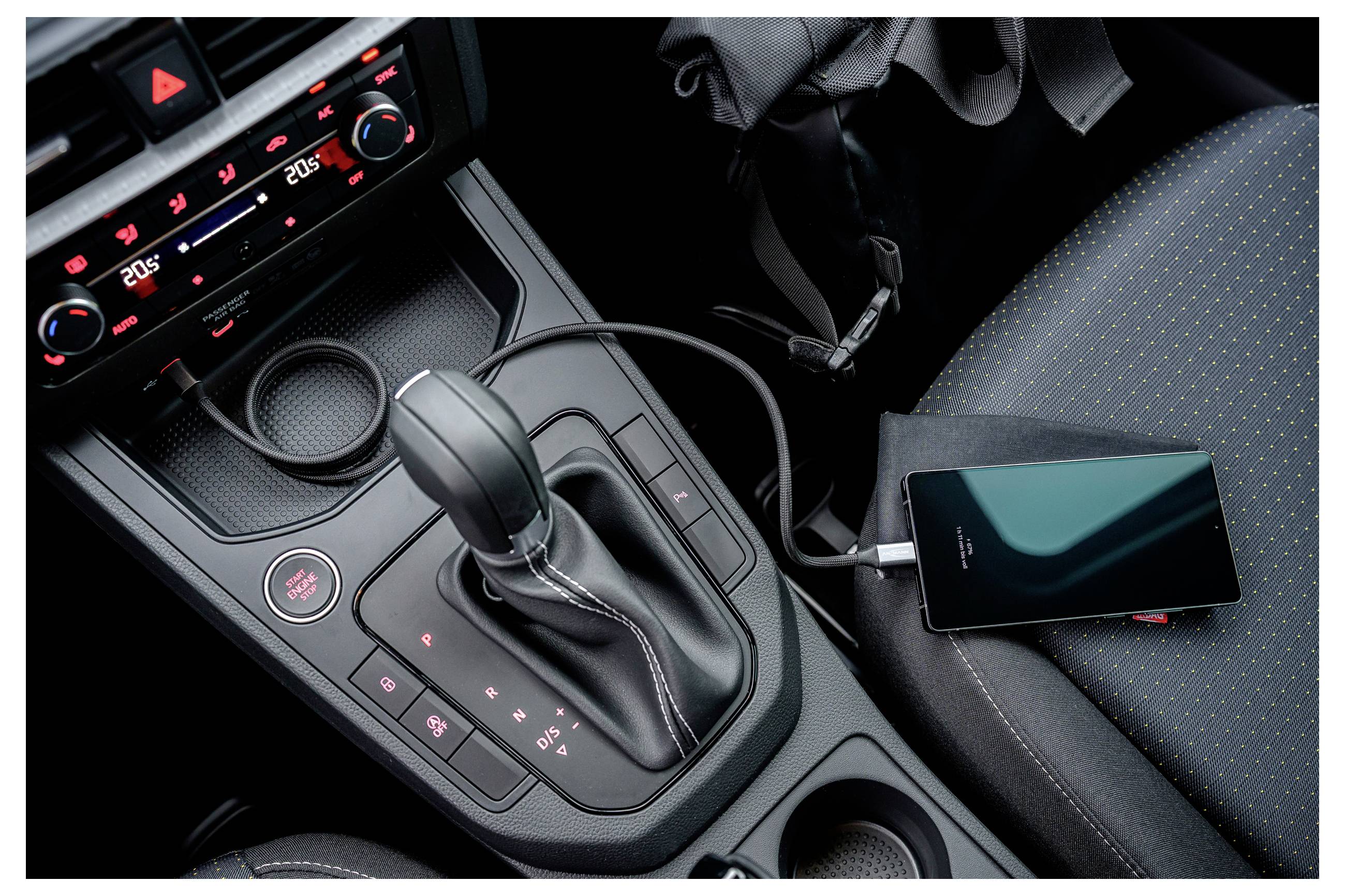 Ein Smartphone, das in einem Auto über ein Kabel aufgeladen wird. Der Schalthebel und die Armaturenbrettsteuerungen des Fahrzeugs sind sichtbar, mit einer Temperaturanzeige.