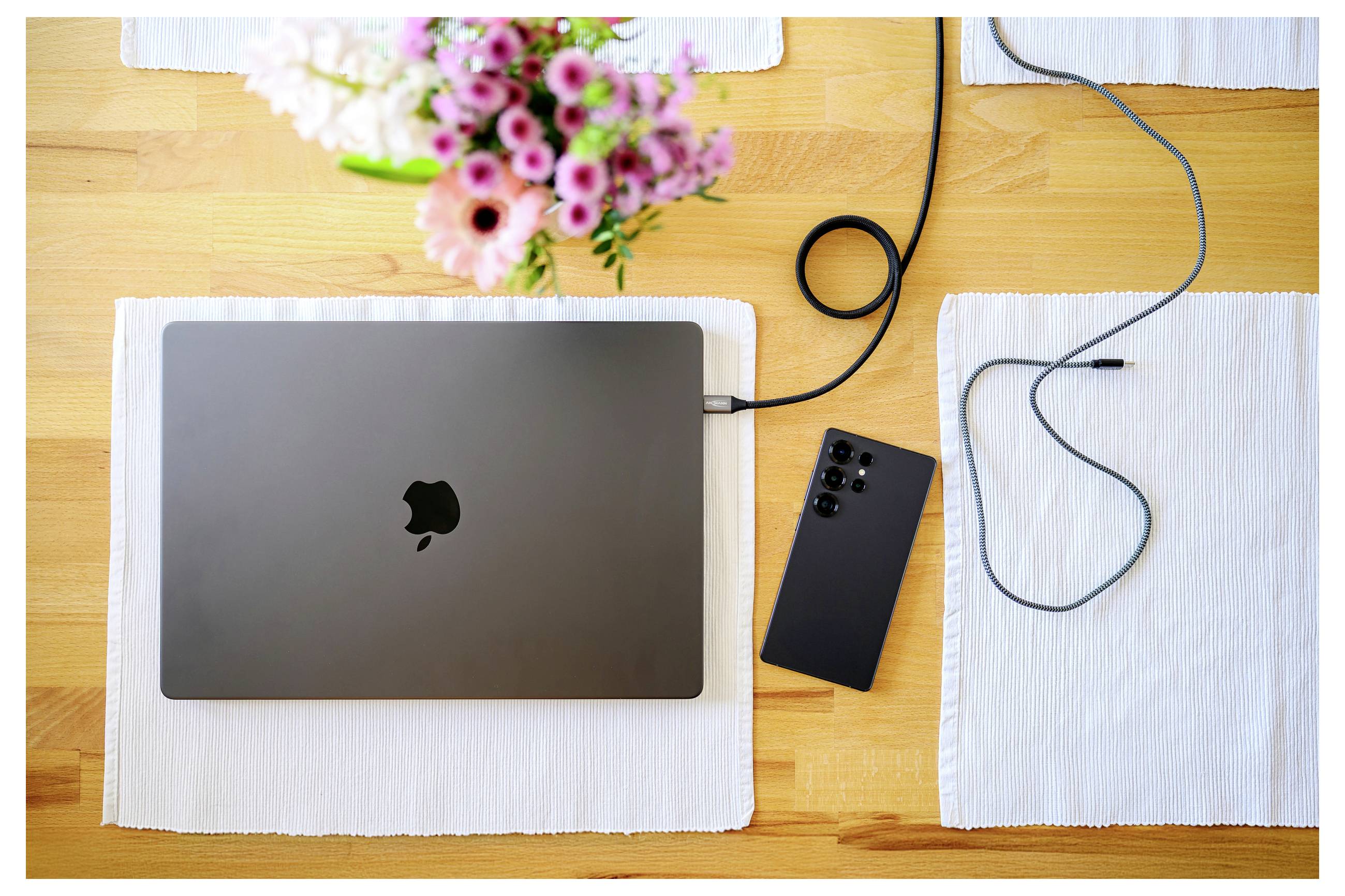 Ein Laptop lädt auf einem Holztisch, umgeben von einem Smartphone, einem Ladekabel und einer Vase mit rosa Blumen.