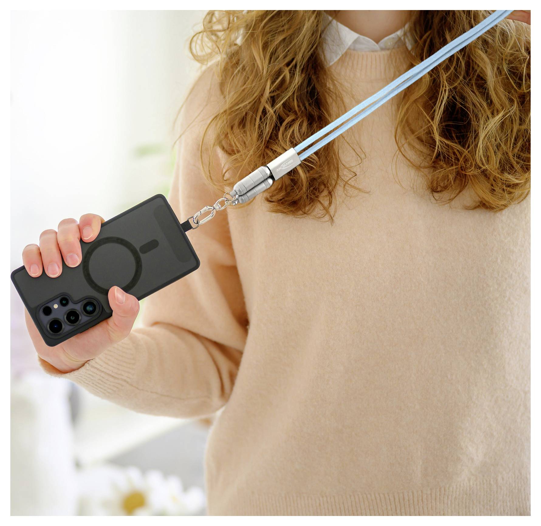 Eine Person trägt einen beigen Pullover und hält ein schwarzes Smartphone, das an einer blauen Lanyard befestigt ist, und demonstriert damit die Möglichkeit, es praktisch freihändig zu tragen.