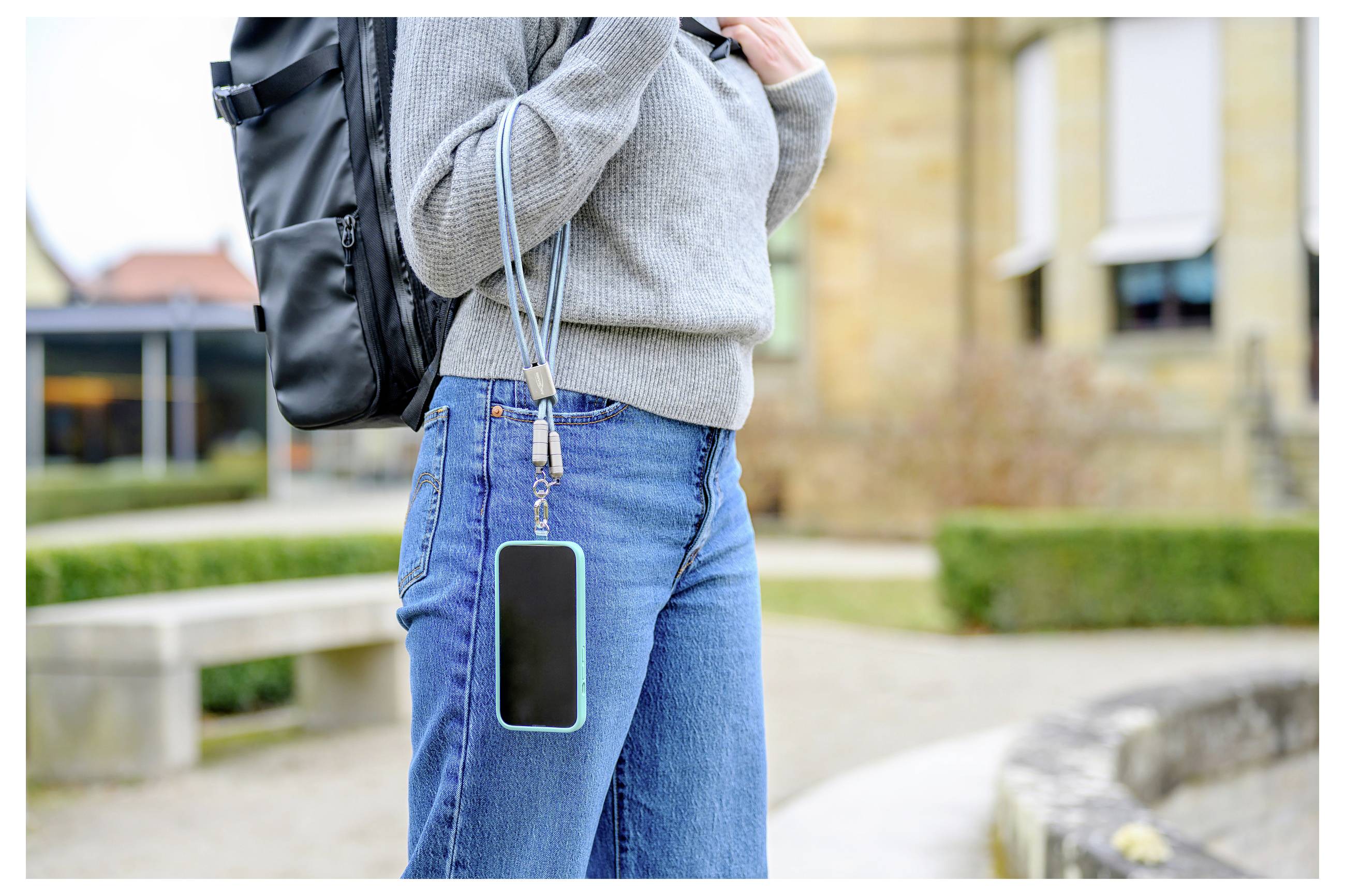Eine Person in einem grauen Pullover trägt einen Rucksack und hat ein Smartphone mit einer Schnur an der Jeans befestigt, während sie sich in einer Outdoor-Umgebung bewegt.