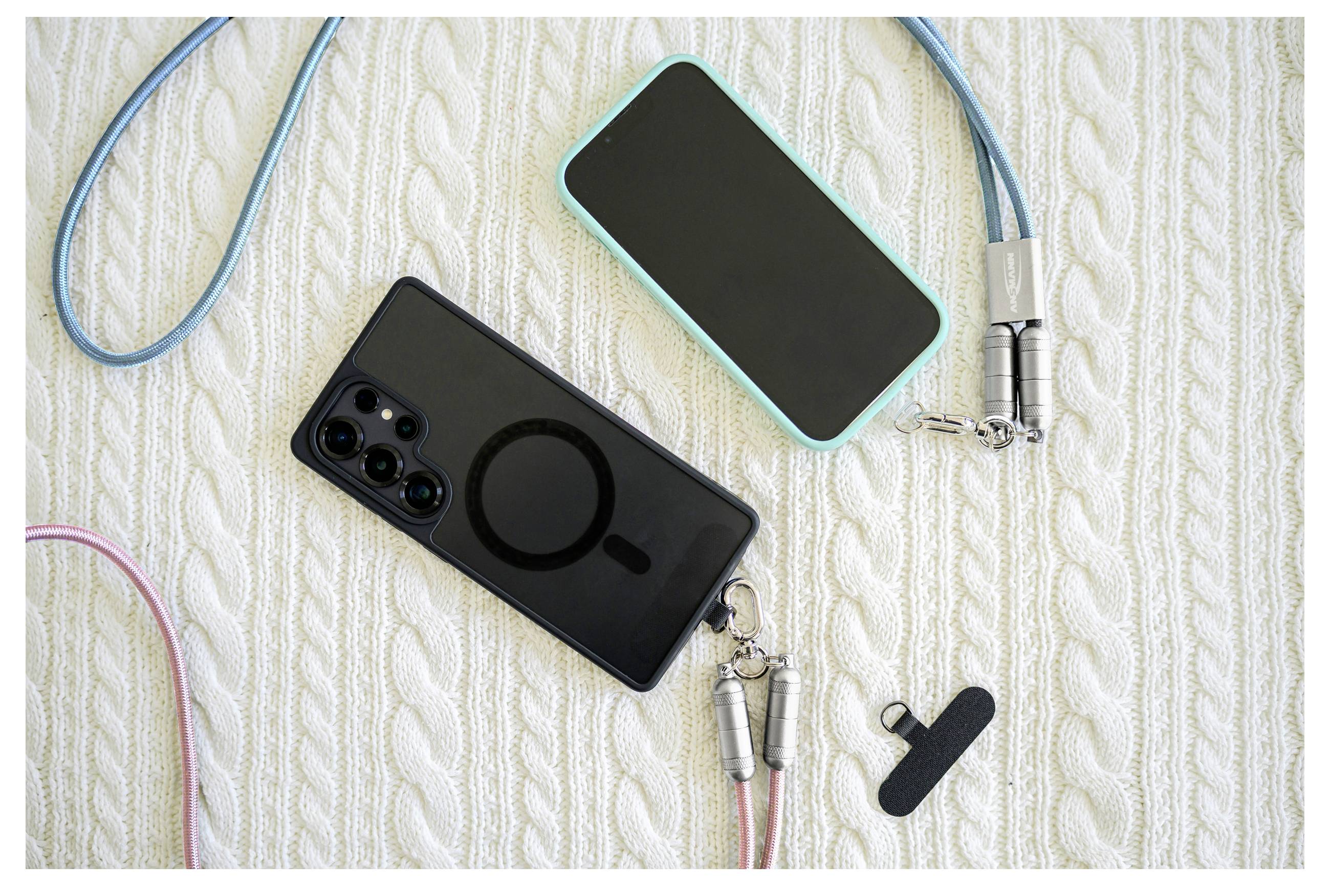 Zwei Smartphones mit Trageband liegen auf einer strukturierten weißen Oberfläche. Ein Telefon ist petrolfarben mit einer vertikalen Kamera, das andere schwarz mit einem Ringdesign.