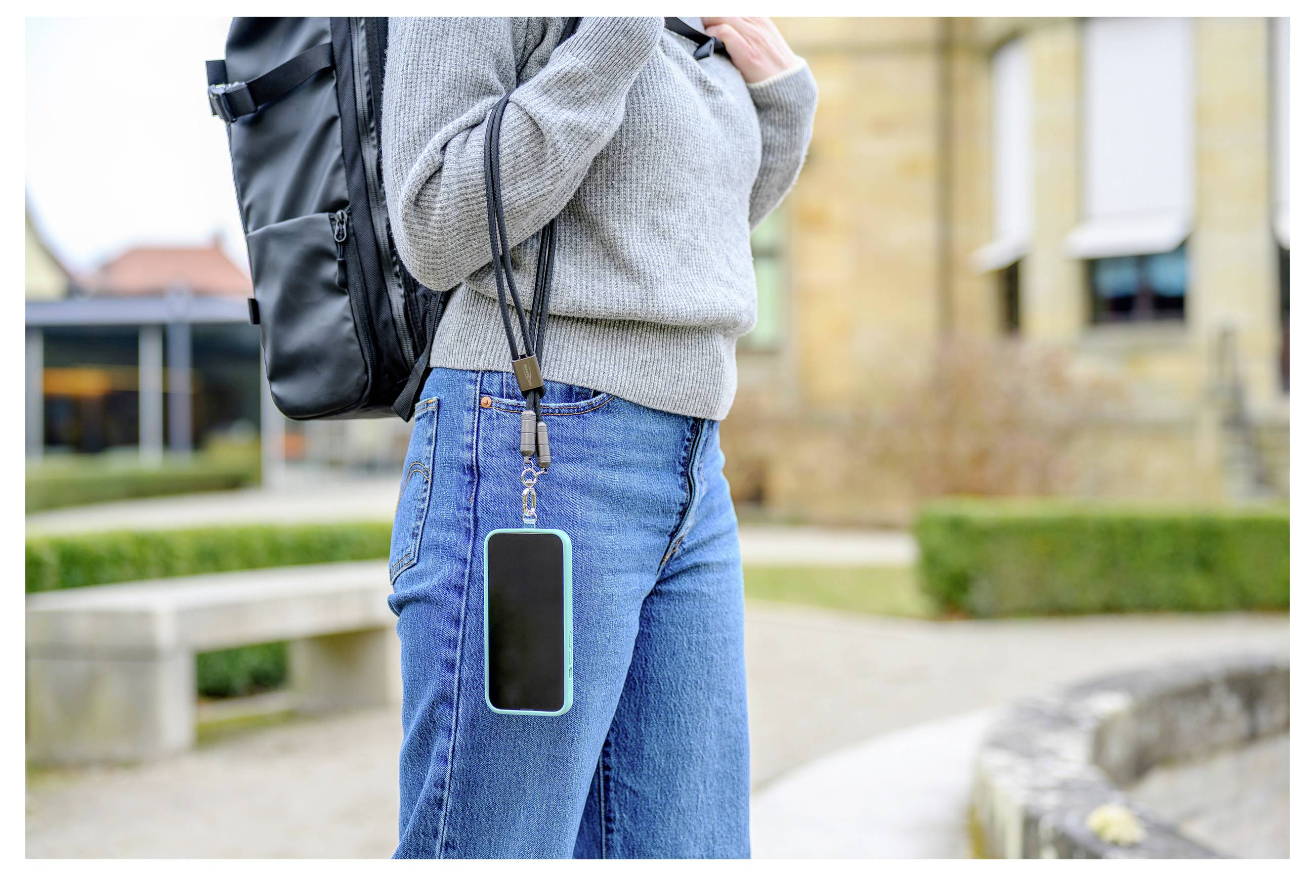 Eine Person im Freien, die einen grauen Pullover und Jeans trägt und einen schwarzen Rucksack bei sich hat. An ihrem Gürtel ist ein Smartphone mit einem Riemen befestigt.