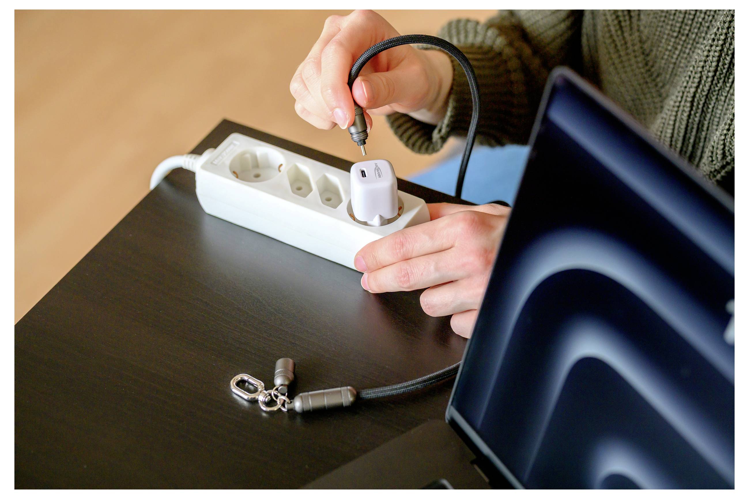 Eine Person steckt ein Netzteil in einen weißen Überspannungsschutz auf einem dunklen Tisch, neben einem Laptop und einem Sicherheitskabel.