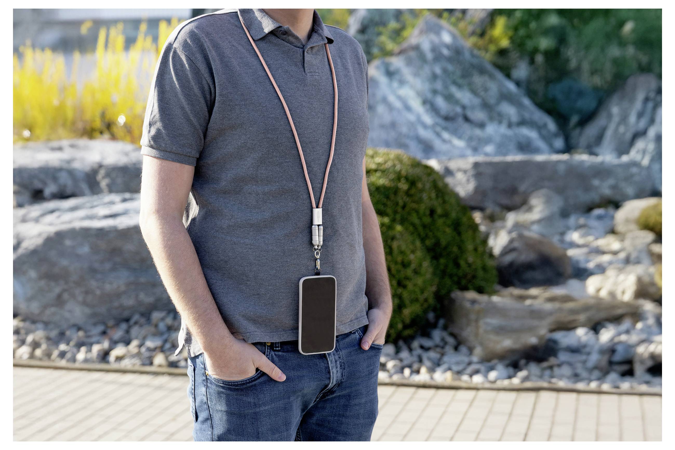 Eine Person steht im Freien, wobei ein Smartphone an einer Lanyard um den Hals hängt, in einer felsigen Gartenlandschaft mit Grünpflanzen.
