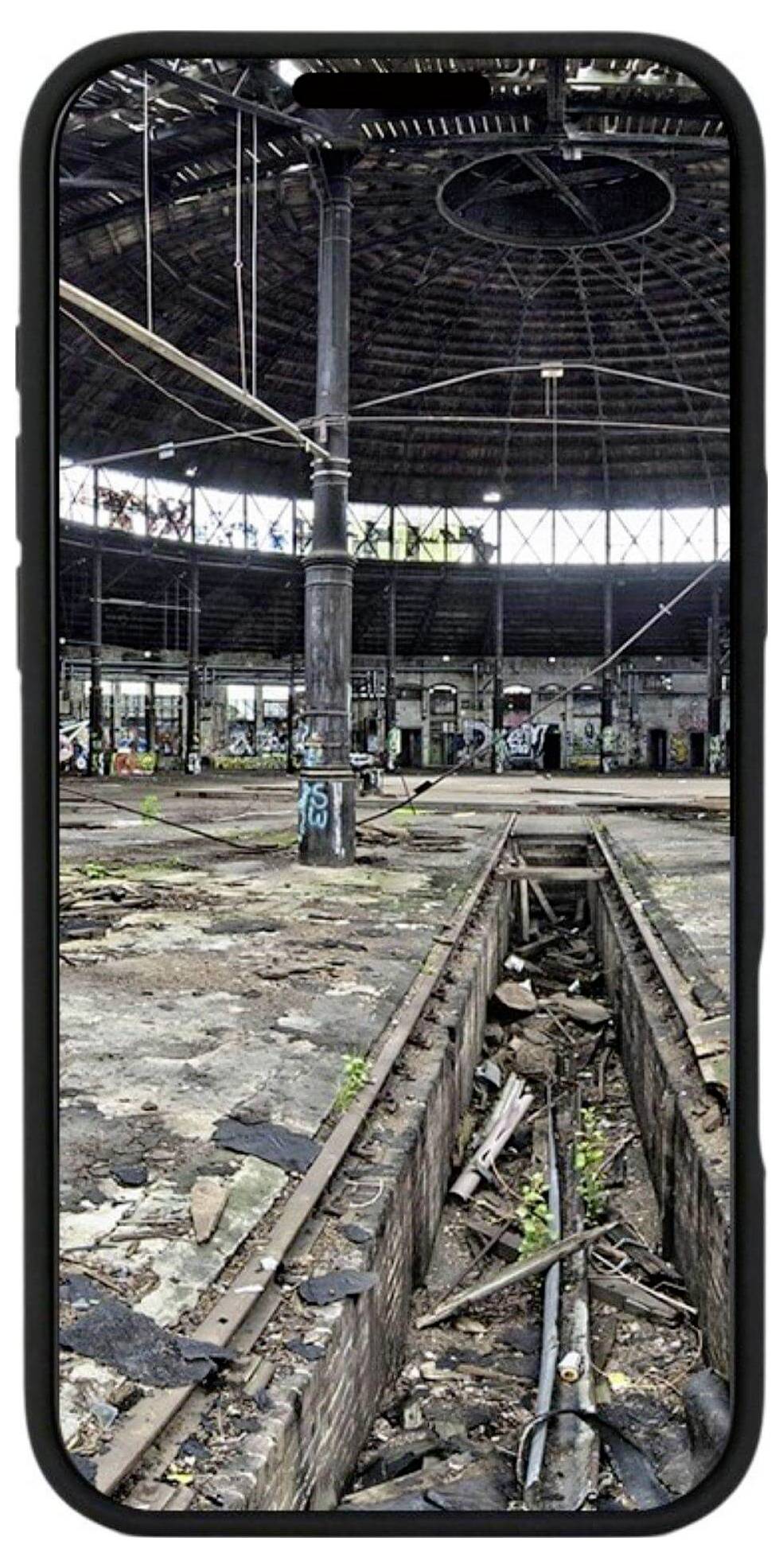 Bild eines verlassenen, mit Graffiti bedeckten Industrieraums, aufgenommen durch einen Smartphone-Bildschirm, mit zerbrochenen Schienen und Trümmern im Inneren.