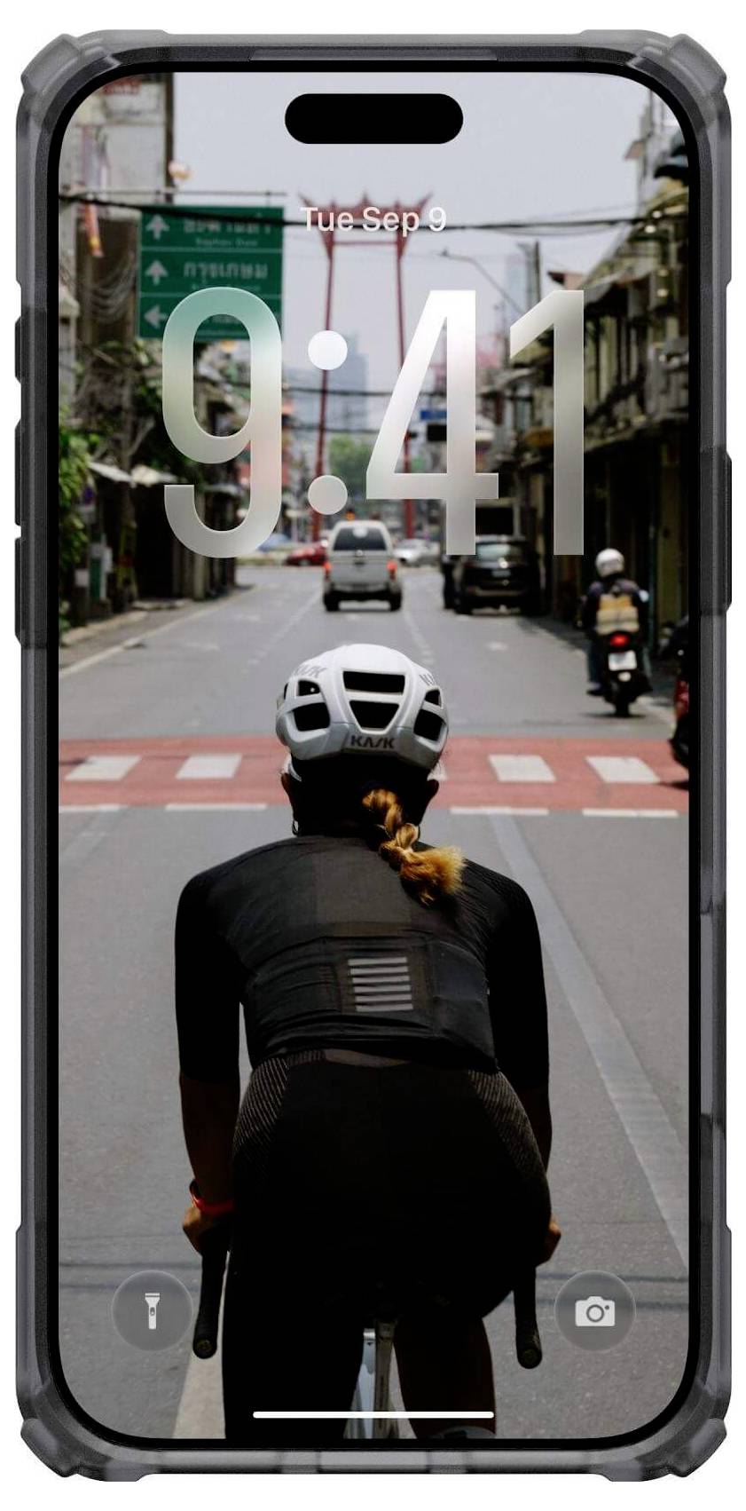 Ein Radfahrer mit Helm fährt auf einen roten Fußgängerüberweg zu, bei klarem Himmel und Gebäuden entlang der Straße, gesehen durch einen Smartphone-Bildschirm, der 9:41 Uhr anzeigt.