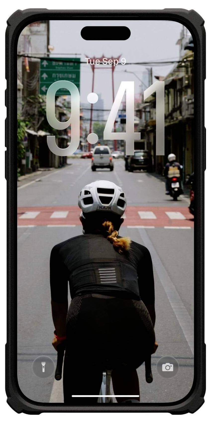Ein Radfahrer mit Helm fährt eine Stadtstraße entlang. Eine digitale Uhr auf einem Handybildschirm zeigt '9:41'. Städtische Gebäude und Fahrzeuge sind zu sehen.