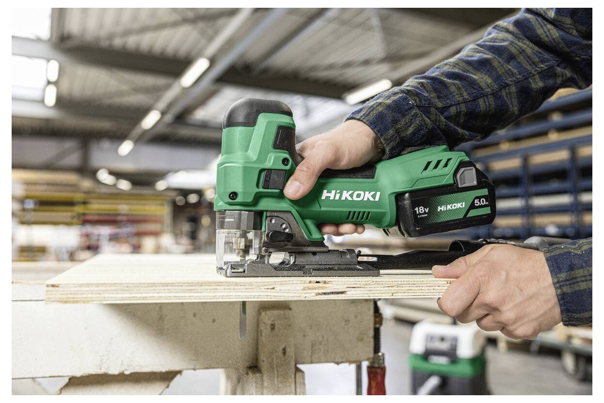 Eine Person verwendet eine grüne Hikoki Stichsäge, um eine Holzplatte in einer gut beleuchteten Werkstatt zu schneiden und dabei deren Präzision und Leistung zu demonstrieren.