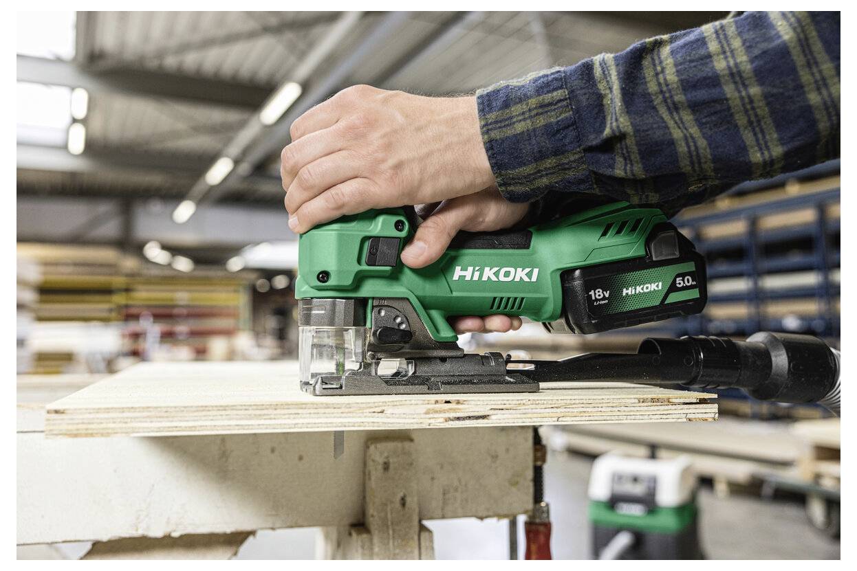 Eine Person verwendet eine grüne Hikoki 18V Stichsäge, um ein Stück Sperrholz in einer Werkstattumgebung zu zersägen.