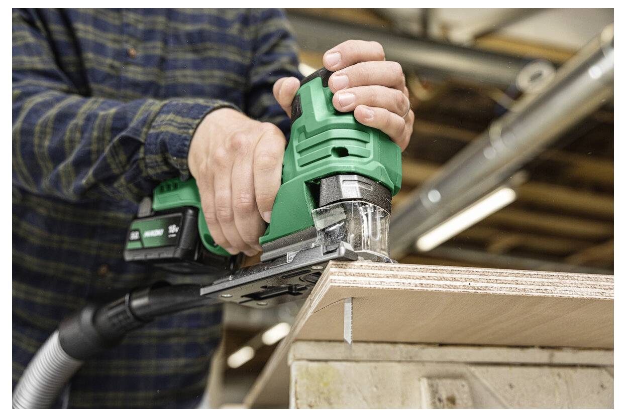 Eine Person, die in einer Werkstatt mit einem Elektrowerkzeug die Kante eines Holzbretts schleift. Das Gerät ist grün und hat einen Staubsammelschlauch angeschlossen.