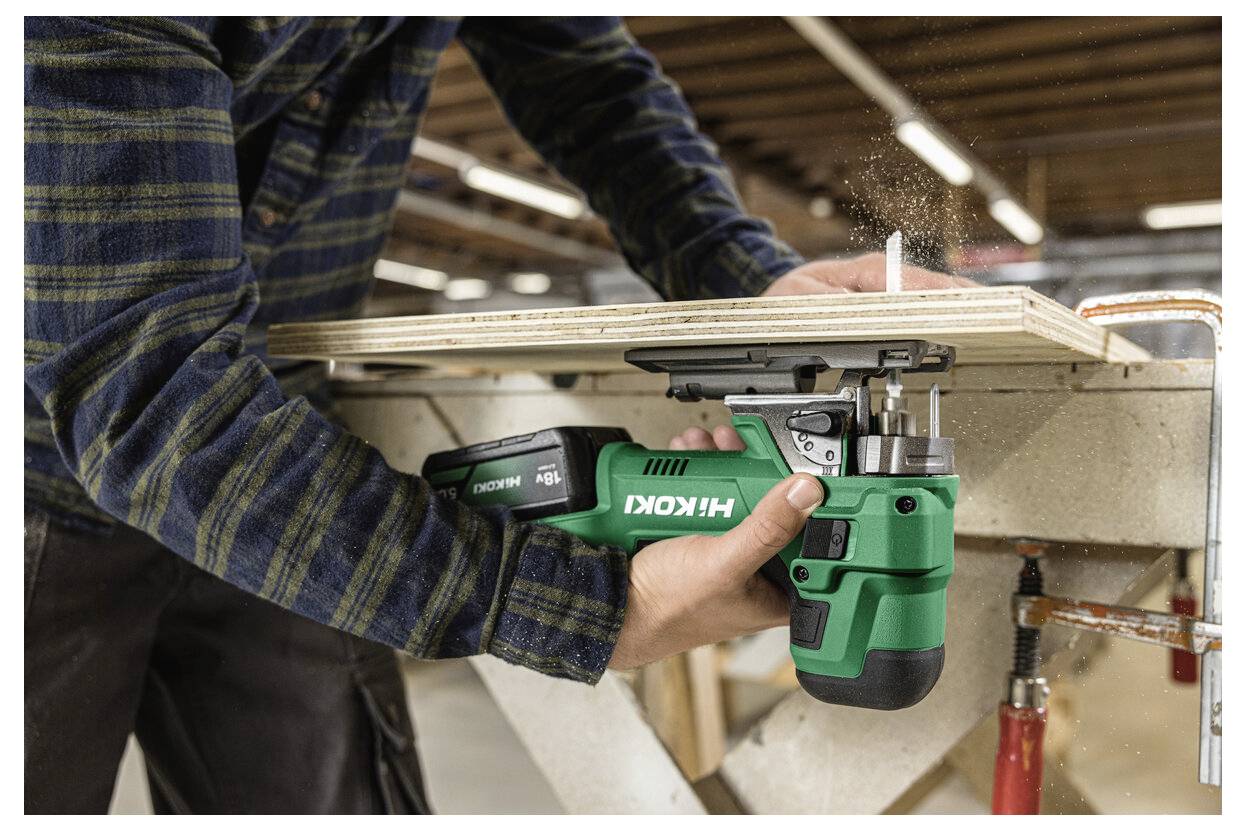 Eine Person verwendet eine grüne Hikoki Akku-Säge, um Sperrholz auf einer Werkbank in einer Werkstatt zu zersägen. Staubpartikel sind in der Luft sichtbar.