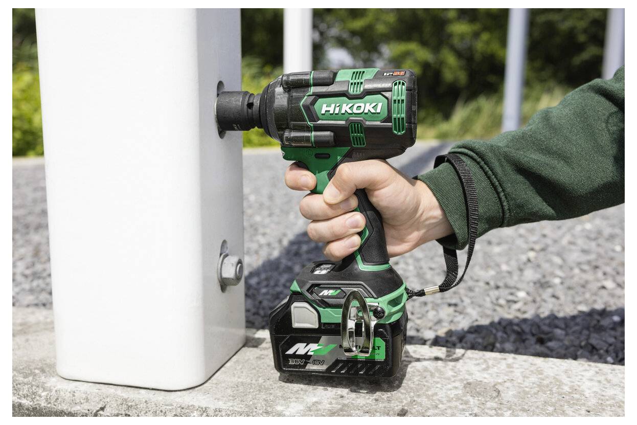 Eine Person verwendet einen kabellosen Schlagschrauber an einem Metallbolzen, der an einem weißen Pfahl im Freien befestigt ist, mit einer Kiesoberfläche und Grünpflanzen im Hintergrund.