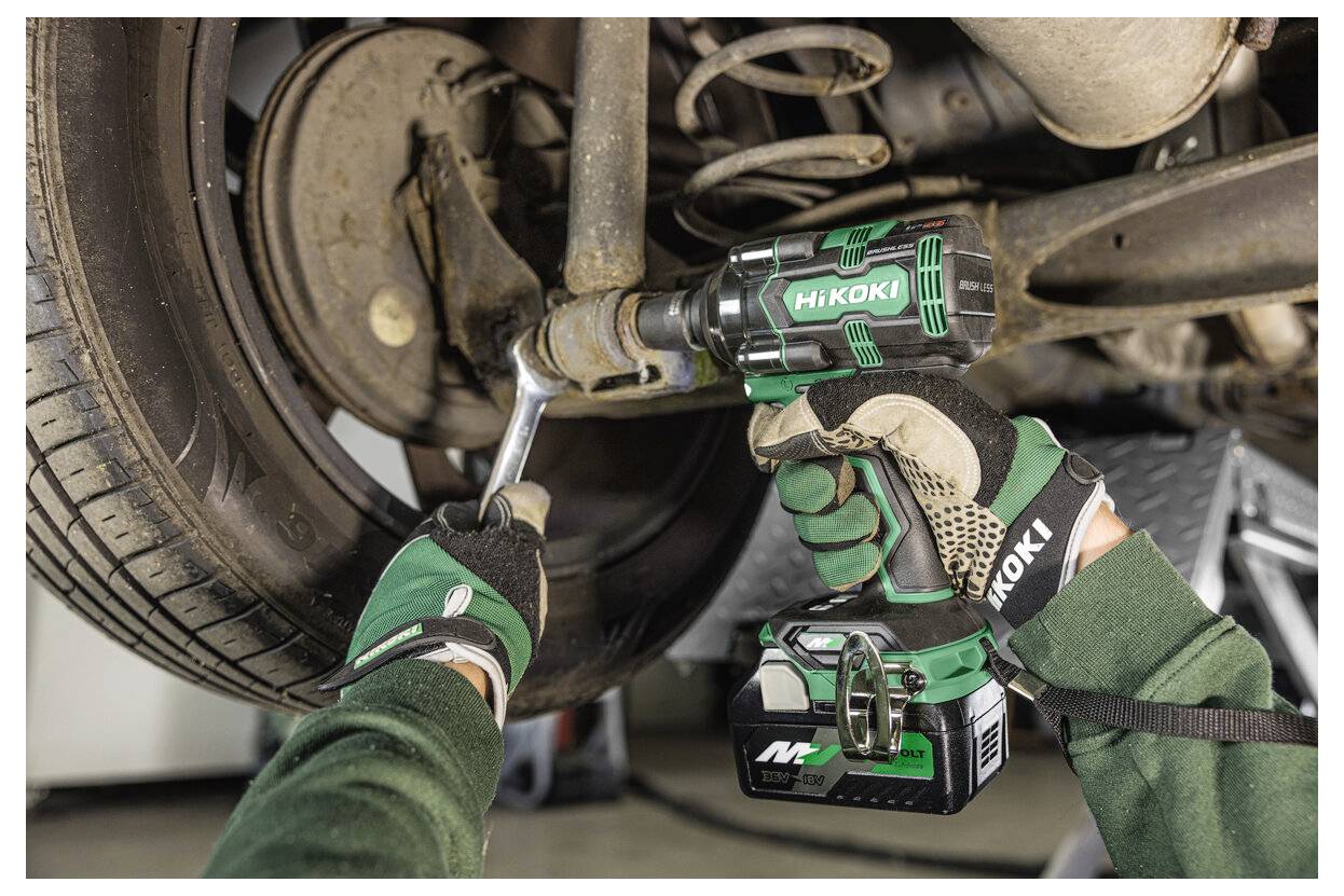 Eine Person in grünen Handschuhen verwendet ein grünes Kraftwerkzeug, um am Fahrzeug-Federsystem zu arbeiten, wobei der Fokus auf einer Radkomponente liegt.