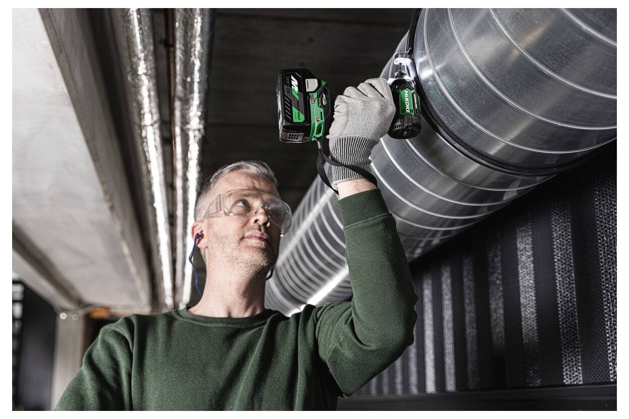 Eine Person mit Sicherheitsbrille und Handschuhen benutzt eine Bohrmaschine, um einen Metallbelüftungskanal in einer industriellen Umgebung zu installieren oder zu reparieren.