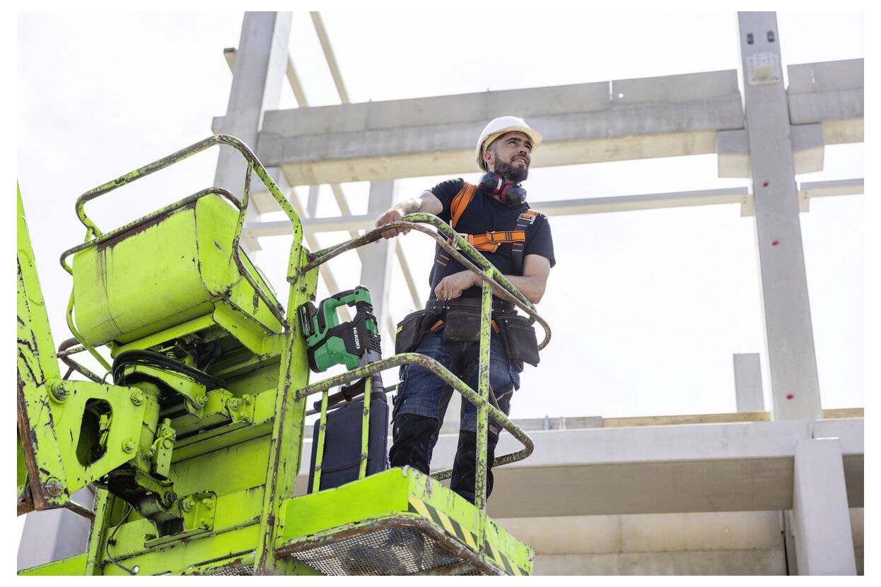 Ein Bauarbeiter in Schutzausrüstung steht auf einer grünen Scherenbühne auf einer Baustelle und beobachtet den Fortschritt des Bauwerks.