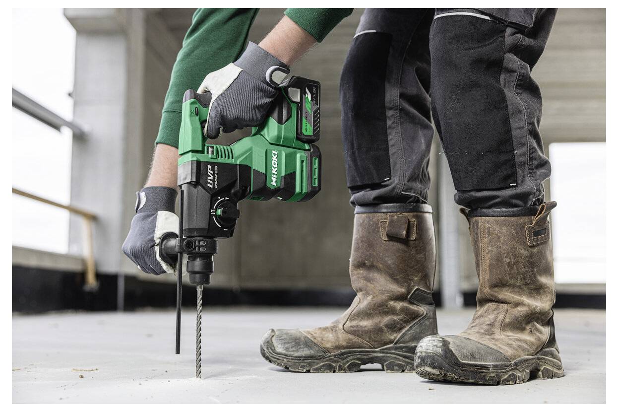 Eine Person mit Handschuhen und Stiefeln verwendet eine grüne Bohrmaschine auf einem Betonboden in einem unfertig Gebäude.
