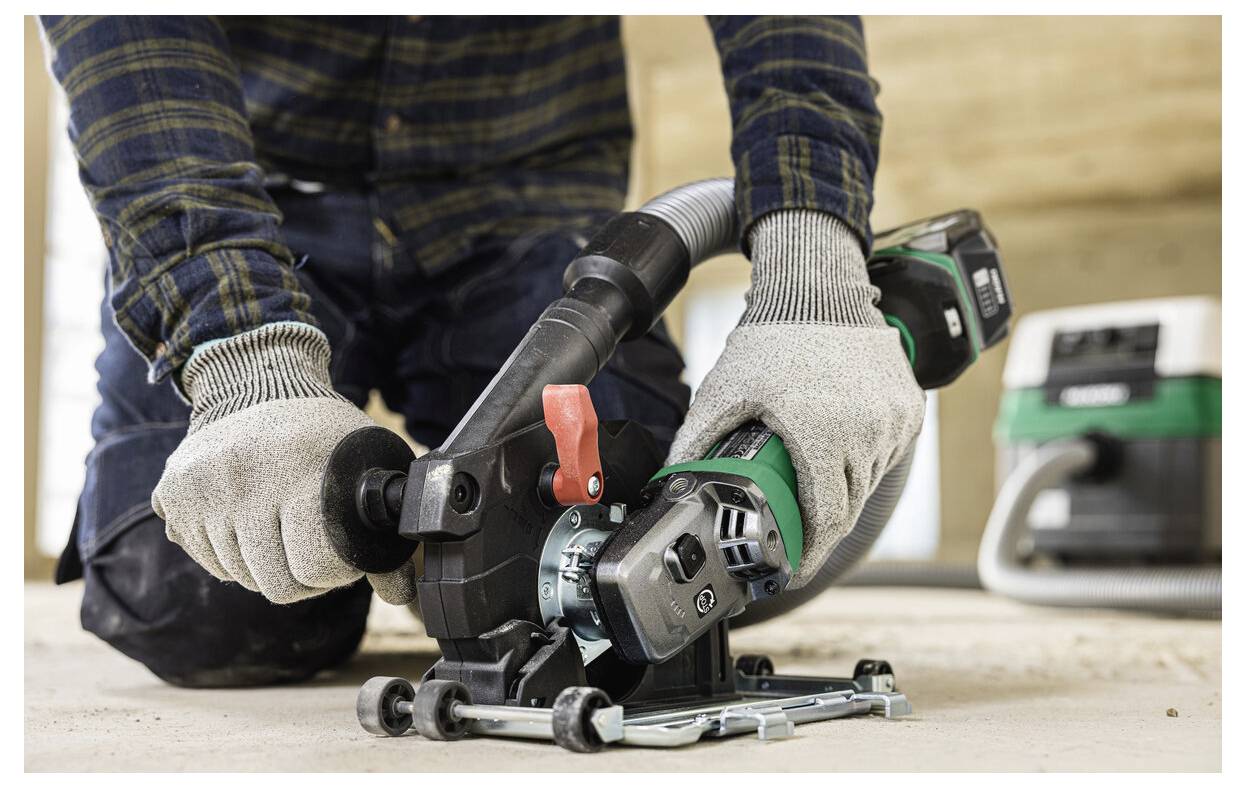 Eine Person mit Handschuhen bedient ein Elektrowerkzeug mit einem Staubsammeladapter. Das Werkzeug wird auf einer Holzoberfläche in einer Baustellenumgebung eingesetzt.